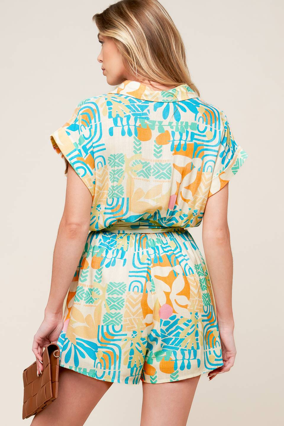 A printed woven romper featuring collar, button down, short cuffed sleeve, elasticized waist and self sash tie.