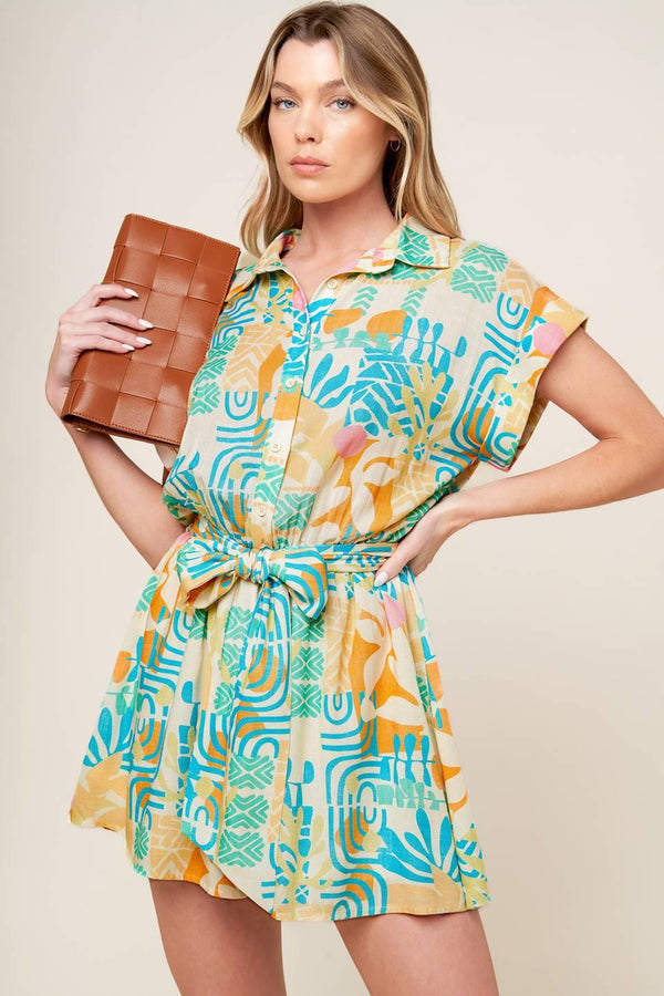 A printed woven romper featuring collar, button down, short cuffed sleeve, elasticized waist and self sash tie.