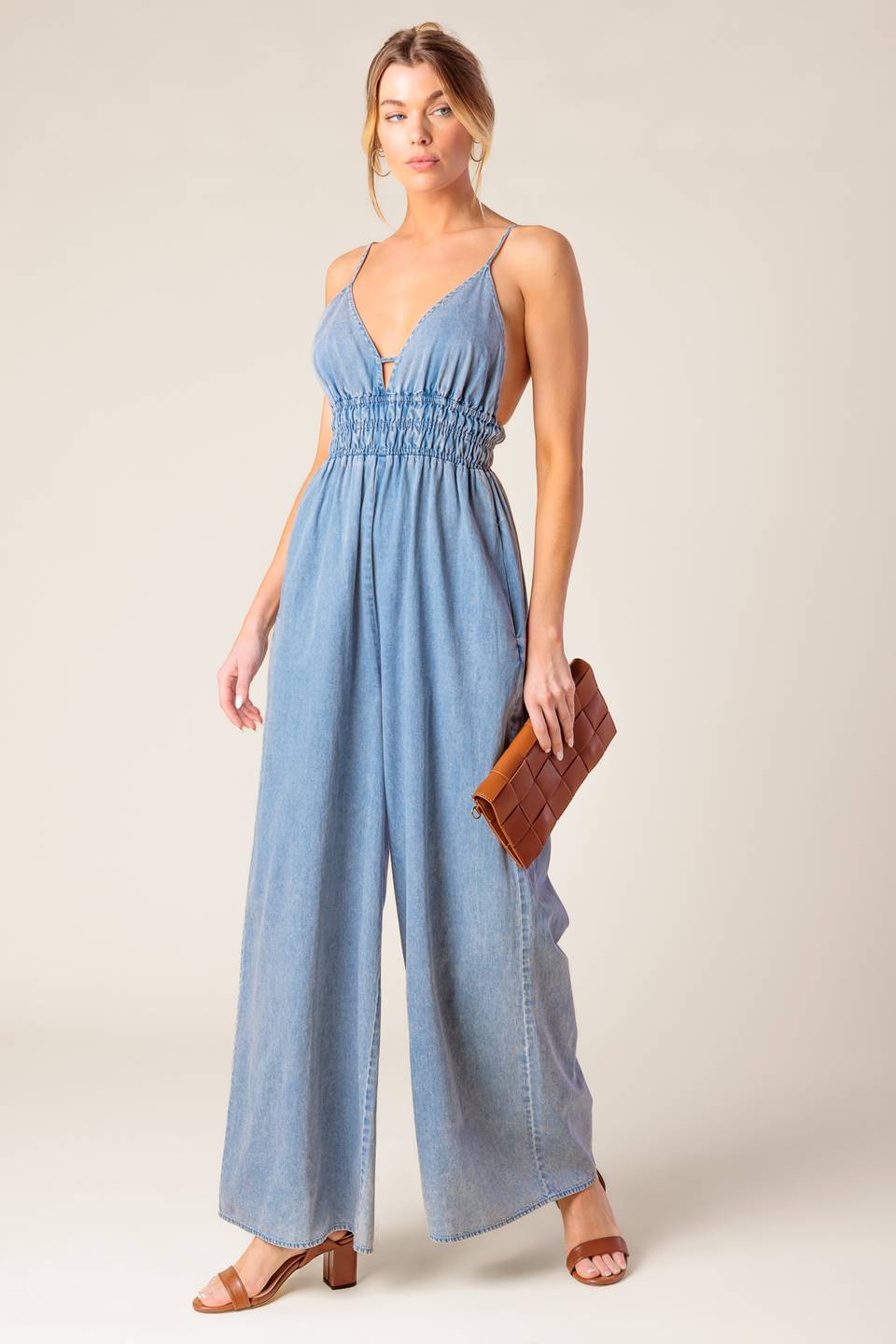 A washed light weight denim jumpsuit featuring V neckline, smocked waist, wide leg and bare back