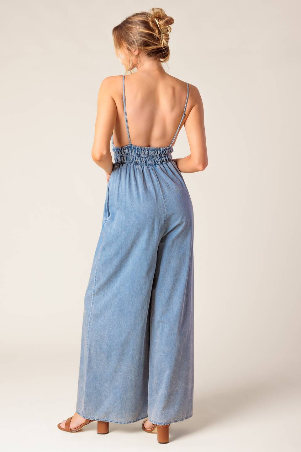 A washed light weight denim jumpsuit featuring V neckline, smocked waist, wide leg and bare back