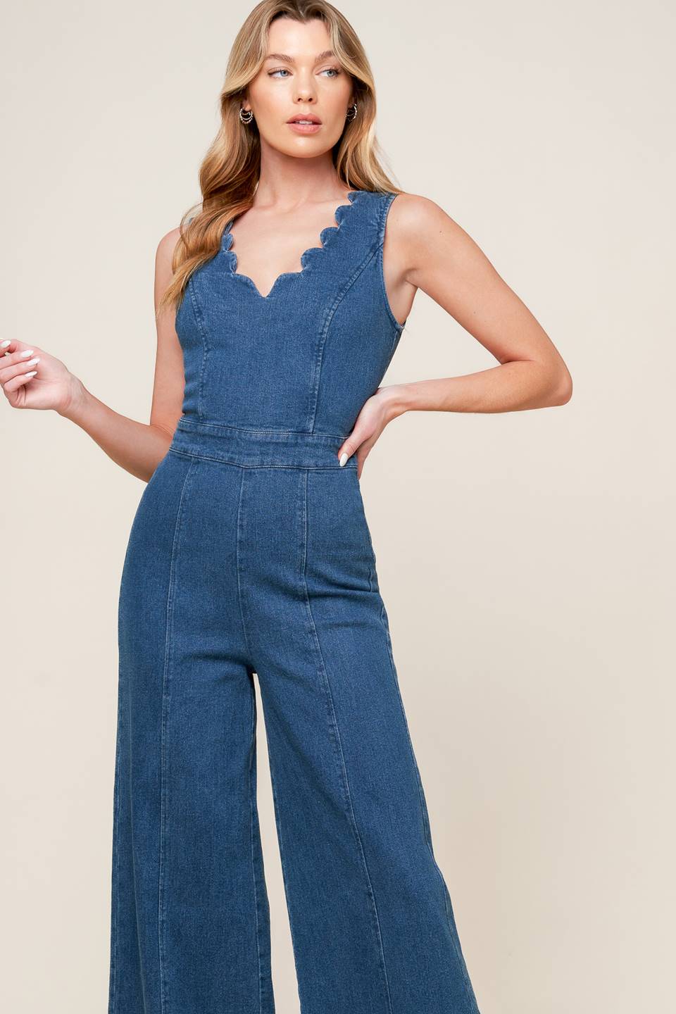 A washed denim jumpsuit featuring scalloped V neckline, sleeveless, self sash belt, wide leg and back zipper closure.