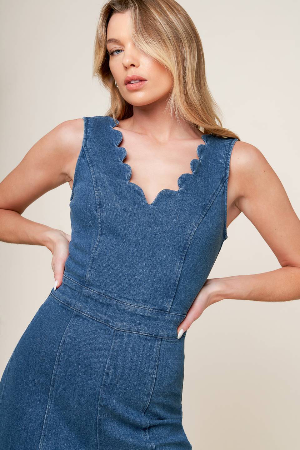 A washed denim jumpsuit featuring scalloped V neckline, sleeveless, self sash belt, wide leg and back zipper closure.