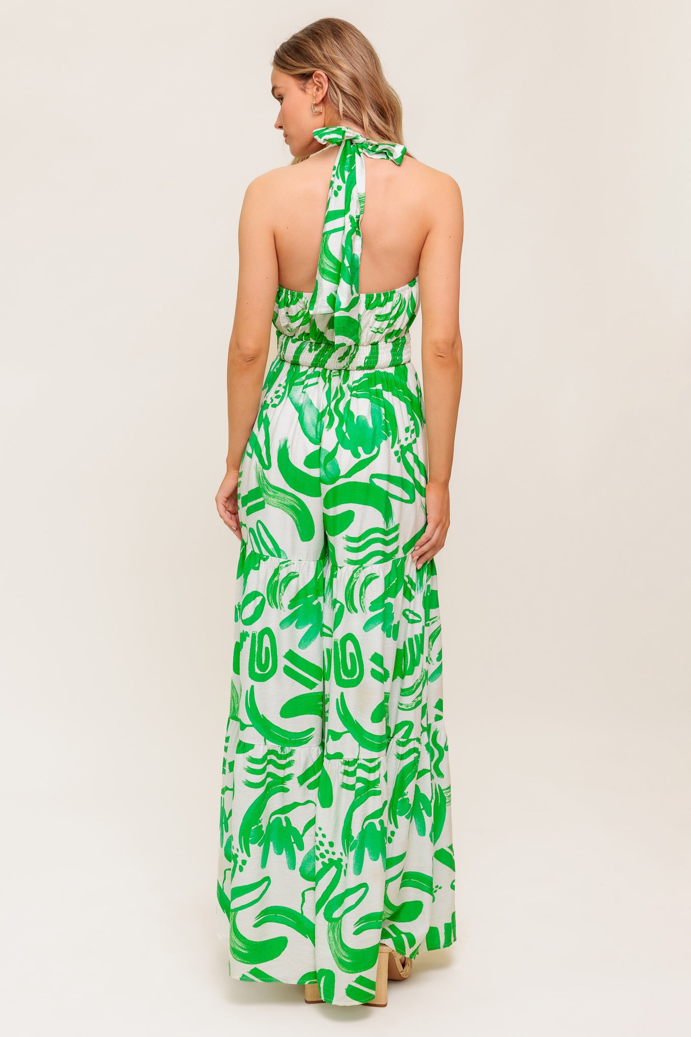 A printed woven jumpsuit featuring halter neckline, tiered pant, bare back with tie and smocked back waistband