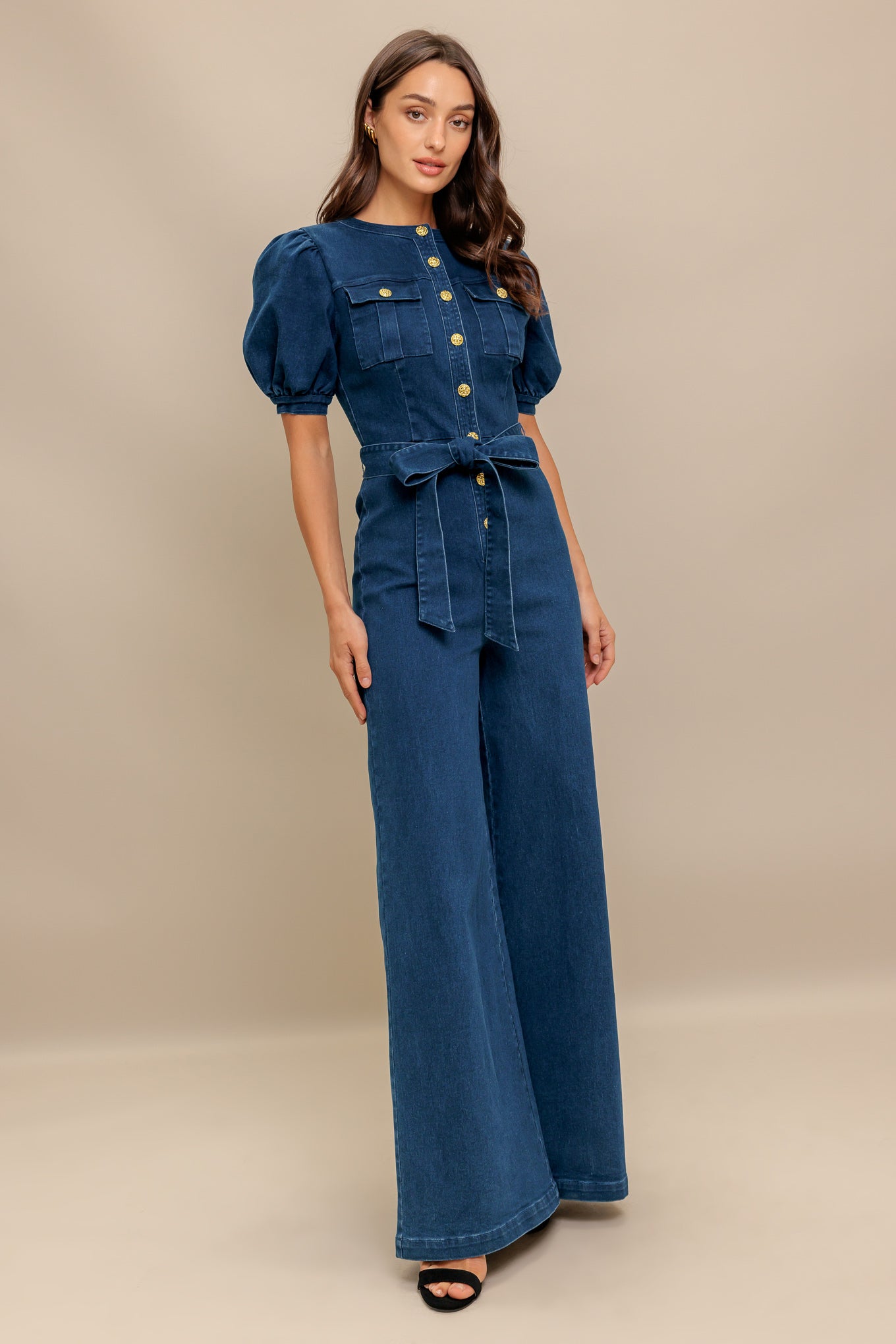 A washed denim jumpsuit featuring round neckline, short puff sleeve, front pockets, button down, self belt and wide leg