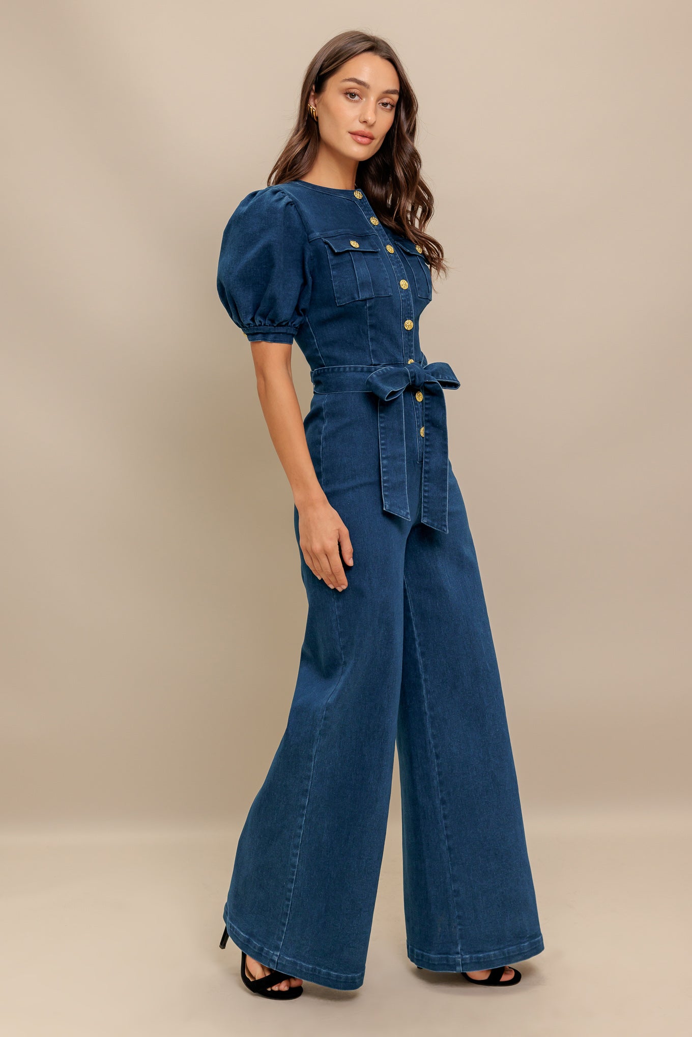 A washed denim jumpsuit featuring round neckline, short puff sleeve, front pockets, button down, self belt and wide leg