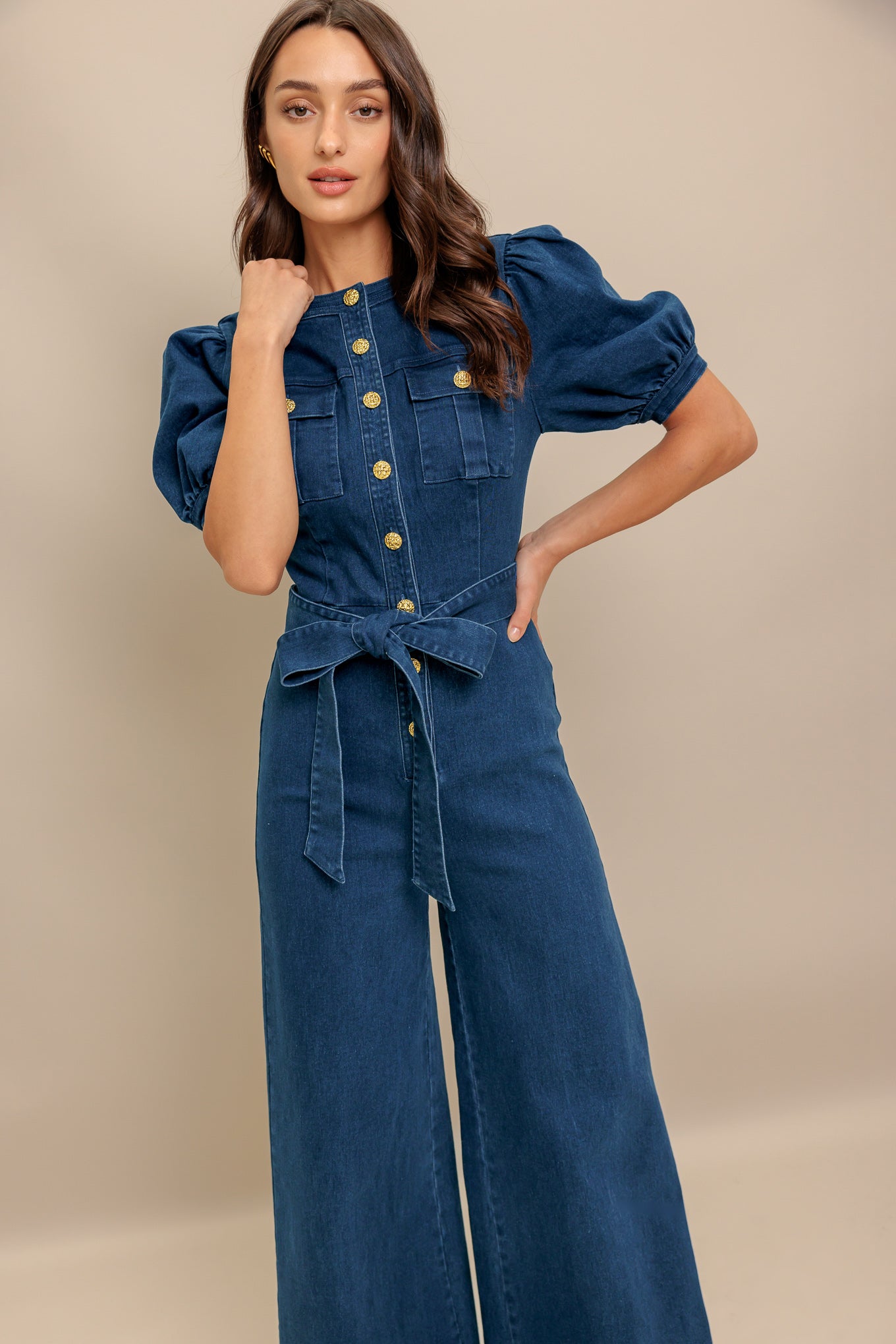 A washed denim jumpsuit featuring round neckline, short puff sleeve, front pockets, button down, self belt and wide leg