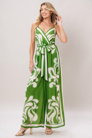 Woman wearing a green and white patterned printed woven jumpsuit featuring surplice neckline, straps, side pockets, self sash tie and wide leg