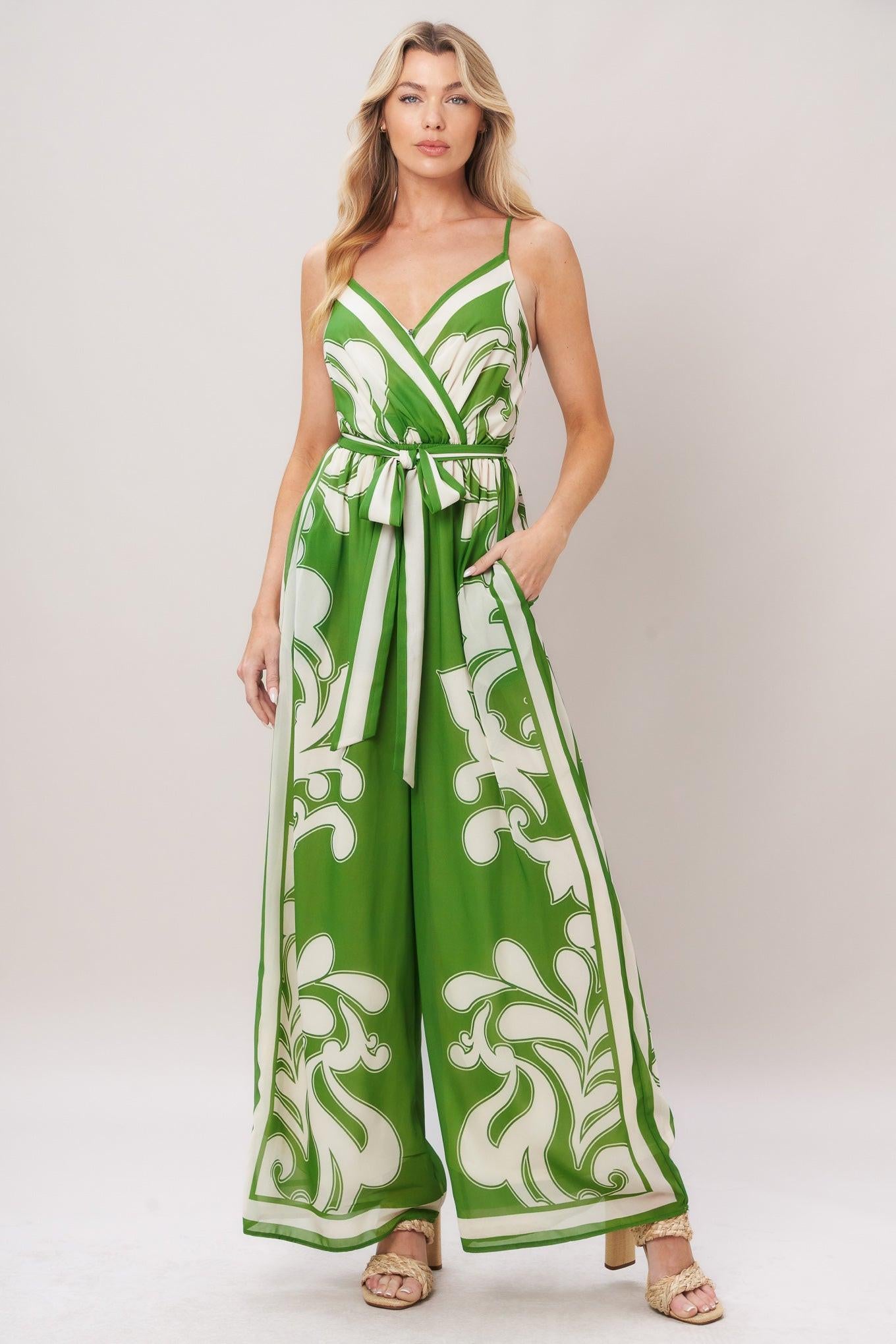 Woman wearing a green and white patterned printed woven jumpsuit featuring surplice neckline, straps, side pockets, self sash tie and wide leg