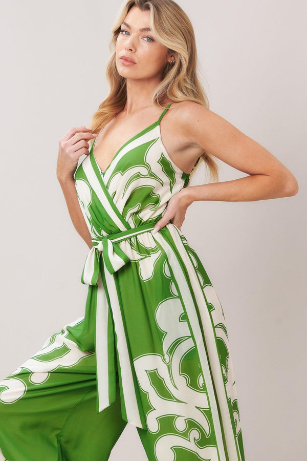 Woman wearing a green and white patterned printed woven jumpsuit featuring surplice neckline, straps, side pockets, self sash tie and wide leg