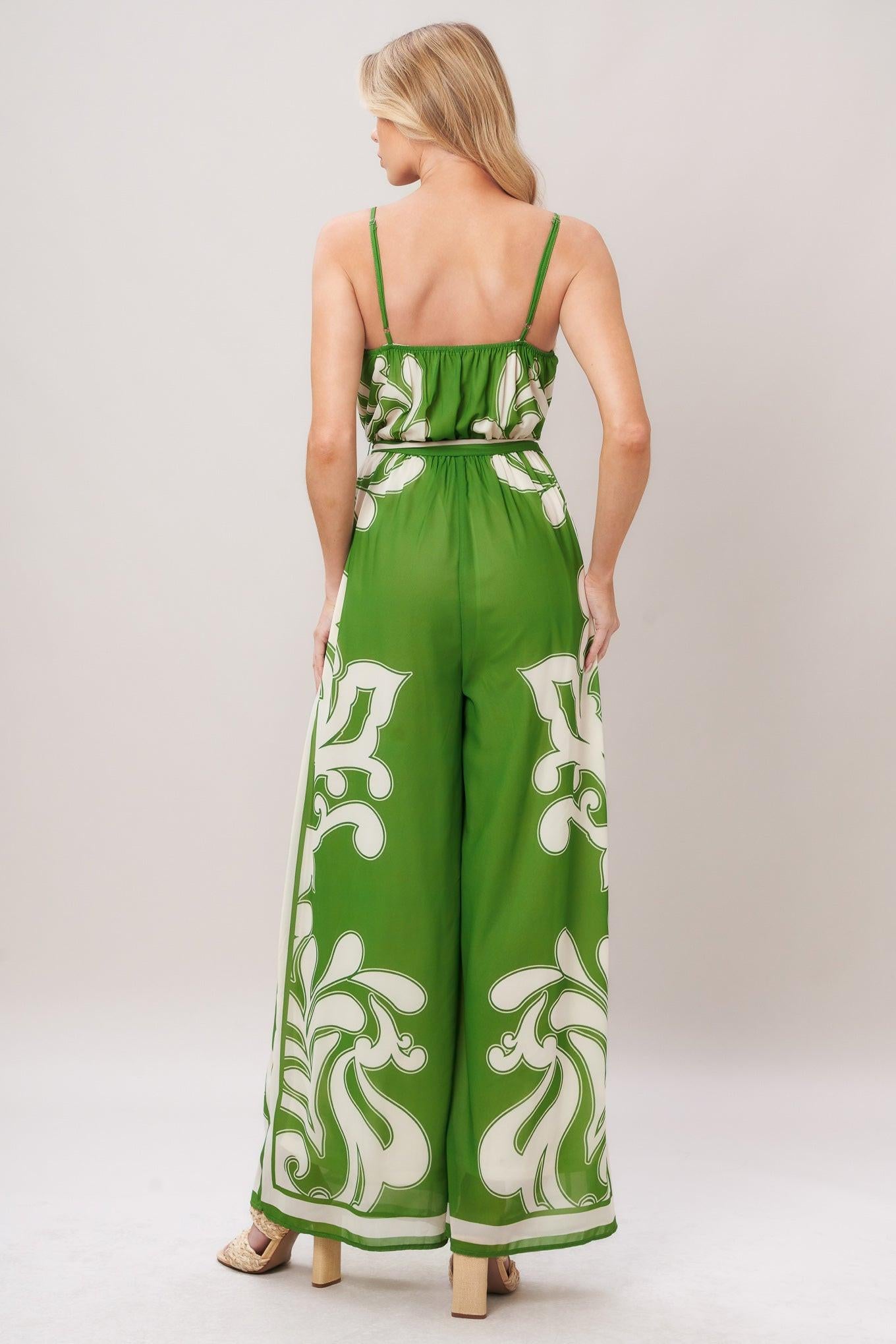 Woman wearing a green and white patterned printed woven jumpsuit featuring surplice neckline, straps, side pockets, self sash tie and wide leg