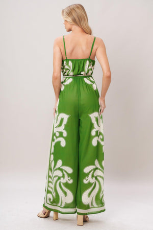 Woman wearing a green and white patterned printed woven jumpsuit featuring surplice neckline, straps, side pockets, self sash tie and wide leg