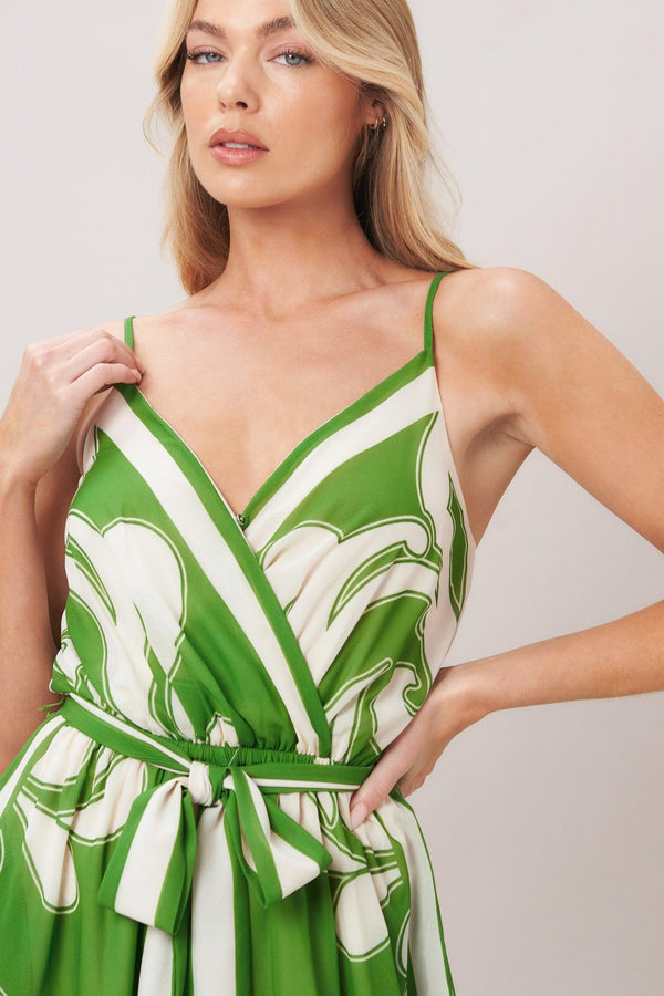 Woman wearing a green and white patterned printed woven jumpsuit featuring surplice neckline, straps, side pockets, self sash tie and wide leg