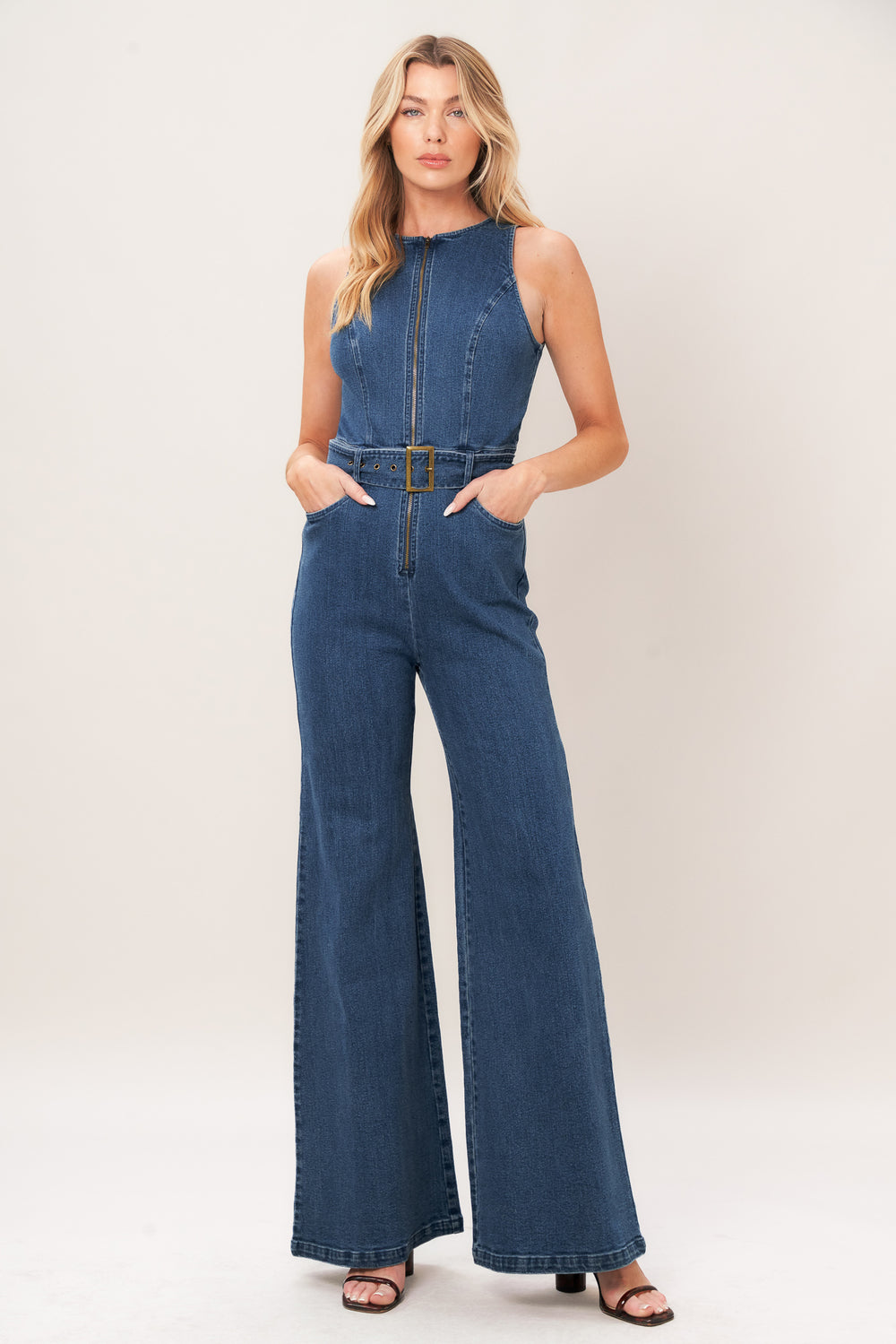 A woman modeling a sleeveless blue denim jumpsuit with wide legs, a front zipper, and a matching belt with a gold-tone buckle, photographed against a plain light beige studio background.