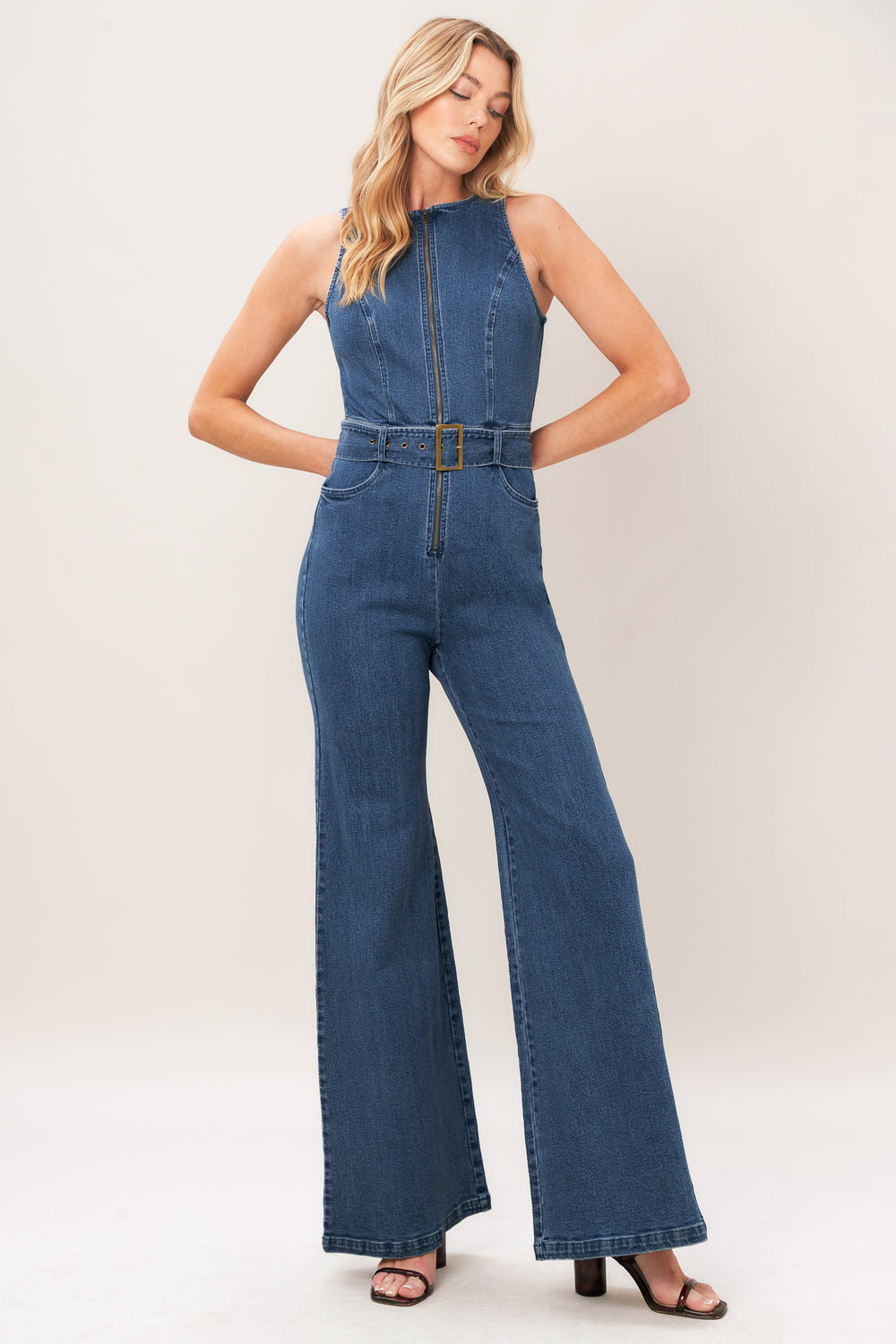 A woman modeling a sleeveless blue denim jumpsuit with wide legs, a front zipper, and a matching belt with a gold-tone buckle, photographed against a plain light beige studio background.