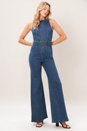 A woman modeling a sleeveless blue denim jumpsuit with wide legs, a front zipper, and a matching belt with a gold-tone buckle, photographed against a plain light beige studio background.
