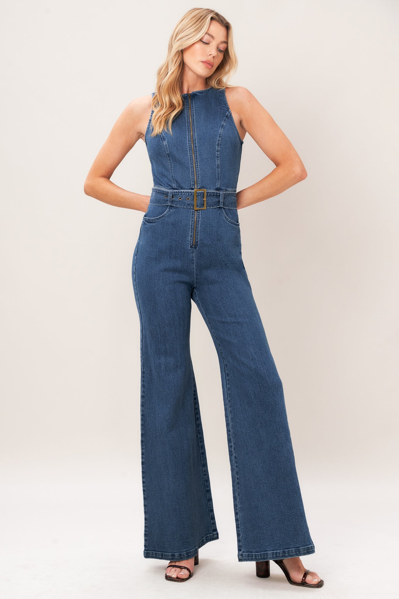 A woman modeling a sleeveless blue denim jumpsuit with wide legs, a front zipper, and a matching belt with a gold-tone buckle, photographed against a plain light beige studio background.