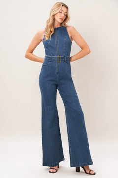 A woman modeling a sleeveless blue denim jumpsuit with wide legs, a front zipper, and a matching belt with a gold-tone buckle, photographed against a plain light beige studio background.
