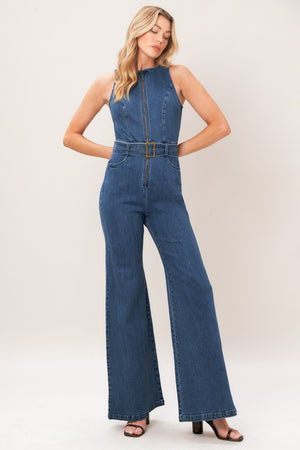 A woman modeling a sleeveless blue denim jumpsuit with wide legs, a front zipper, and a matching belt with a gold-tone buckle, photographed against a plain light beige studio background.