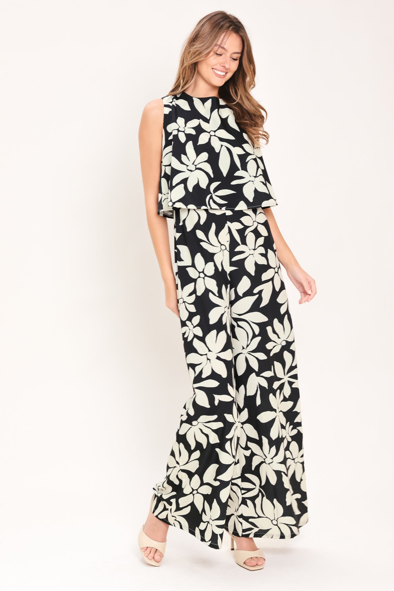 Woman wearing a black and white floral wide leg jumpsuit featuring round neckline, popover, split back with zipper closure | Flying Tomato