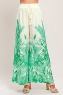 Woman wearing green and white tropical leaf patterned wide-leg pants with elastic waist and front tie | Flying Tomato