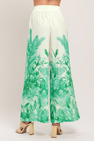 Woman wearing green and white tropical leaf patterned wide-leg pants with elastic waist and front tie | Flying Tomato