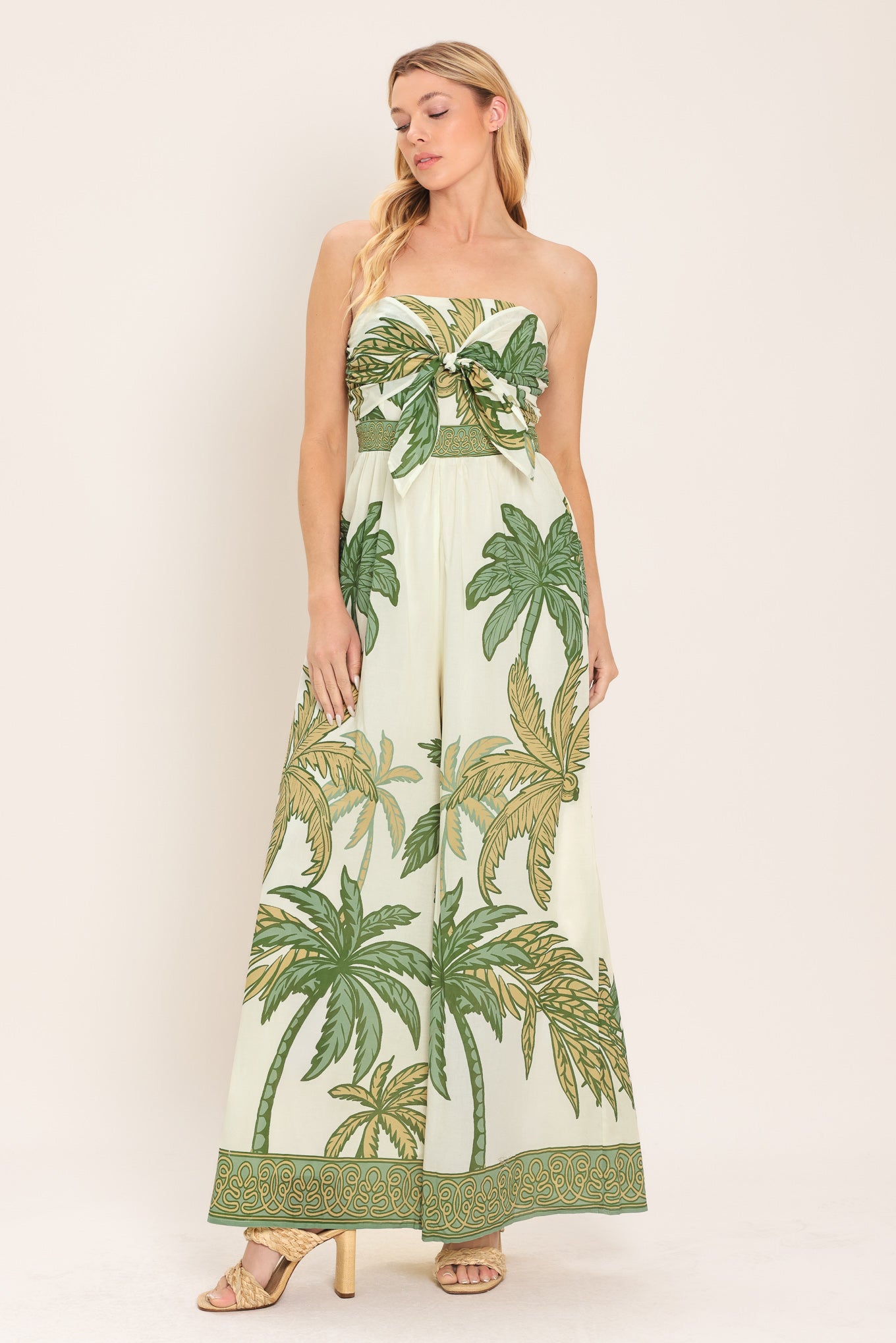 Woman wearing a strapless ivory wide-leg jumpsuit with green palm tree pattern featuring front tie and smocked back bodice | Flying Tomato