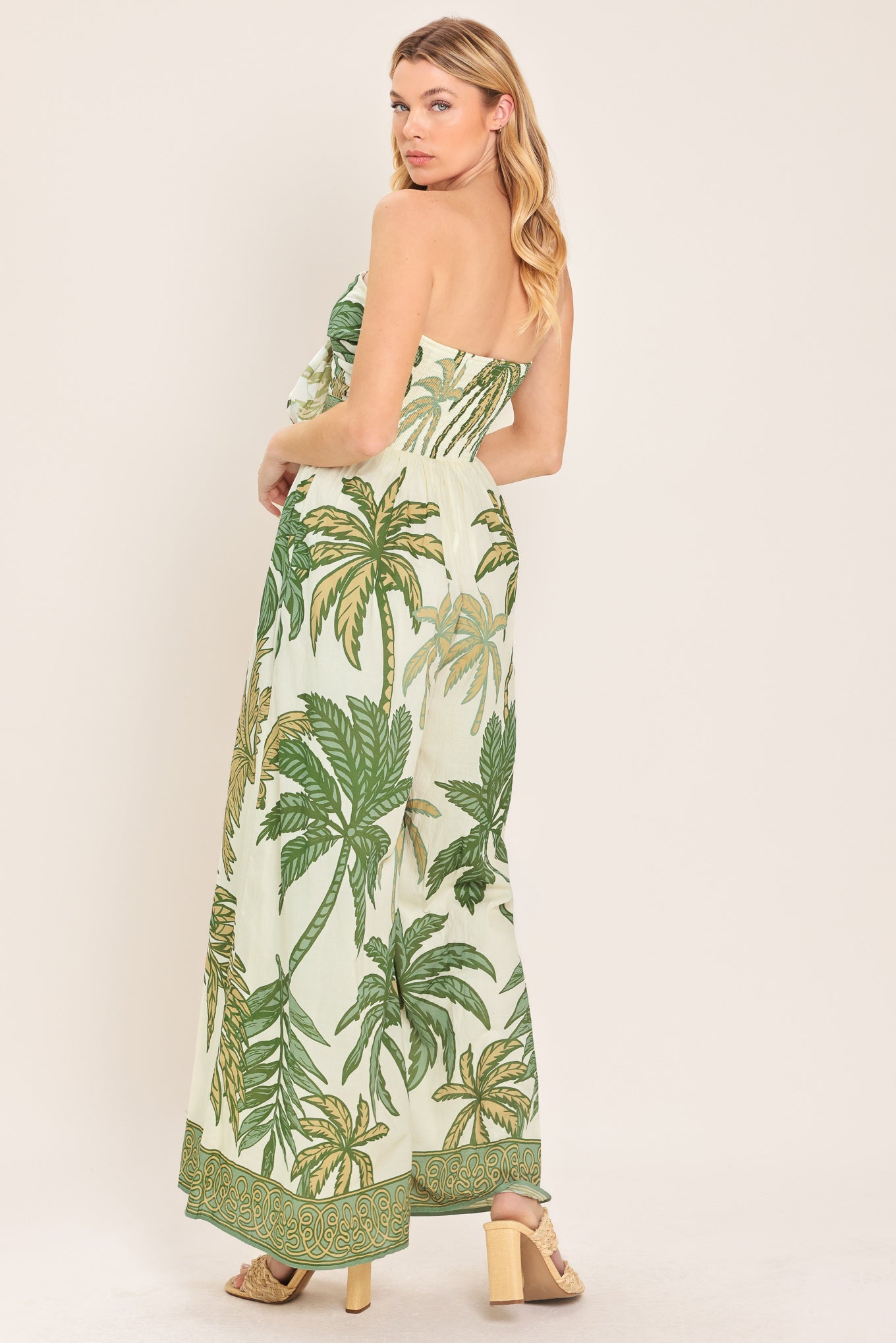 Woman wearing a strapless ivory wide-leg jumpsuit with green palm tree pattern featuring front tie and smocked back bodice | Flying Tomato