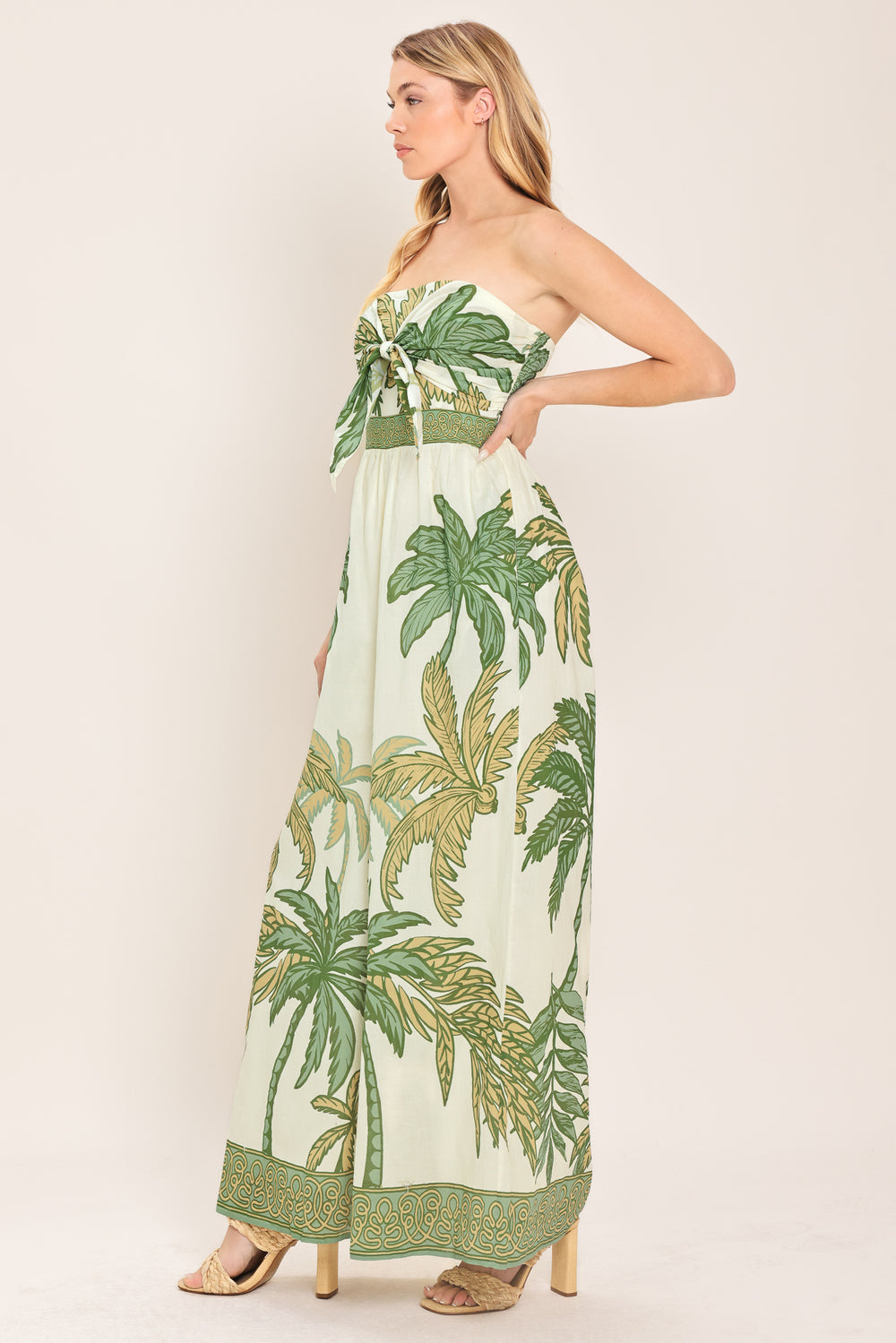 Woman wearing a strapless ivory wide-leg jumpsuit with green palm tree pattern featuring front tie and smocked back bodice | Flying Tomato
