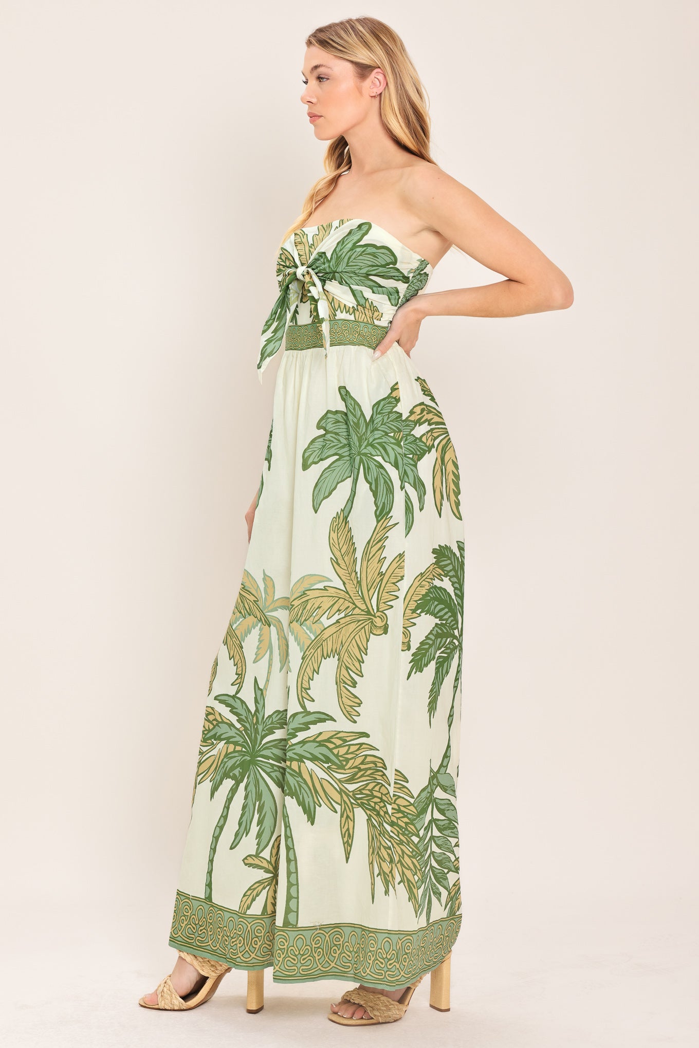 Woman wearing a strapless ivory wide-leg jumpsuit with green palm tree pattern featuring front tie and smocked back bodice | Flying Tomato