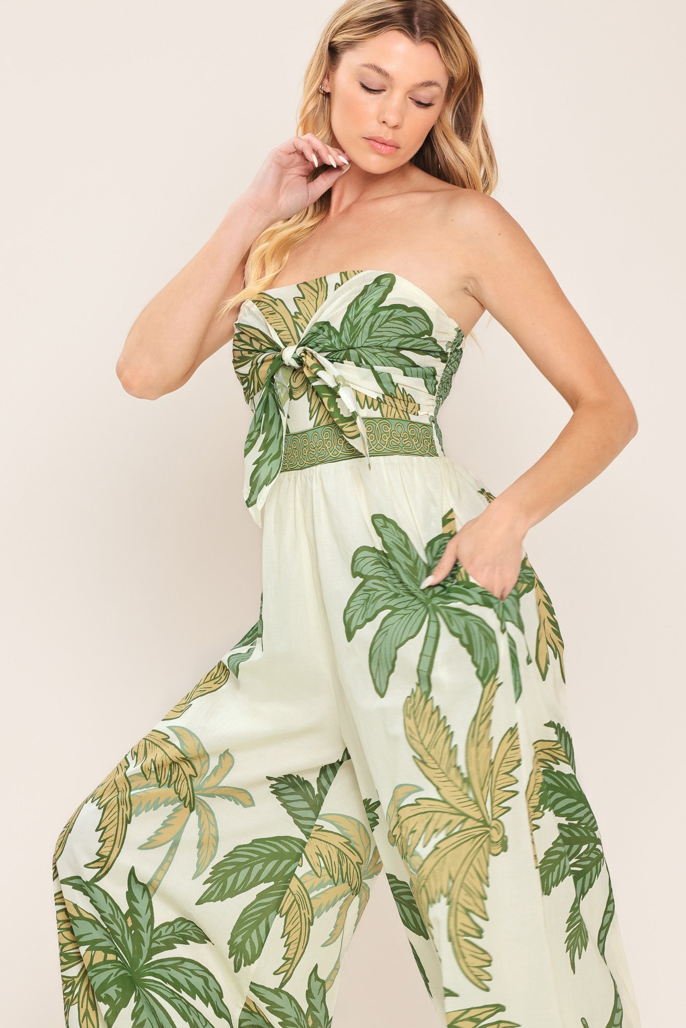 Woman wearing a strapless ivory wide-leg jumpsuit with green palm tree pattern featuring front tie and smocked back bodice | Flying Tomato