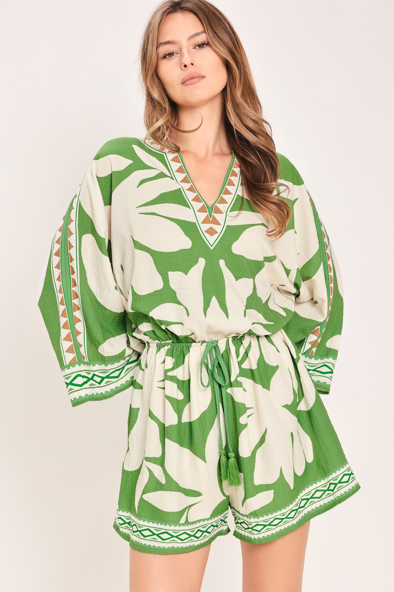 Woman wearing a green and white leaf pattern romper featuring V neckline, kimono sleeve, elastic waist with tie tassel and back neck button closure | Flying Tomato
