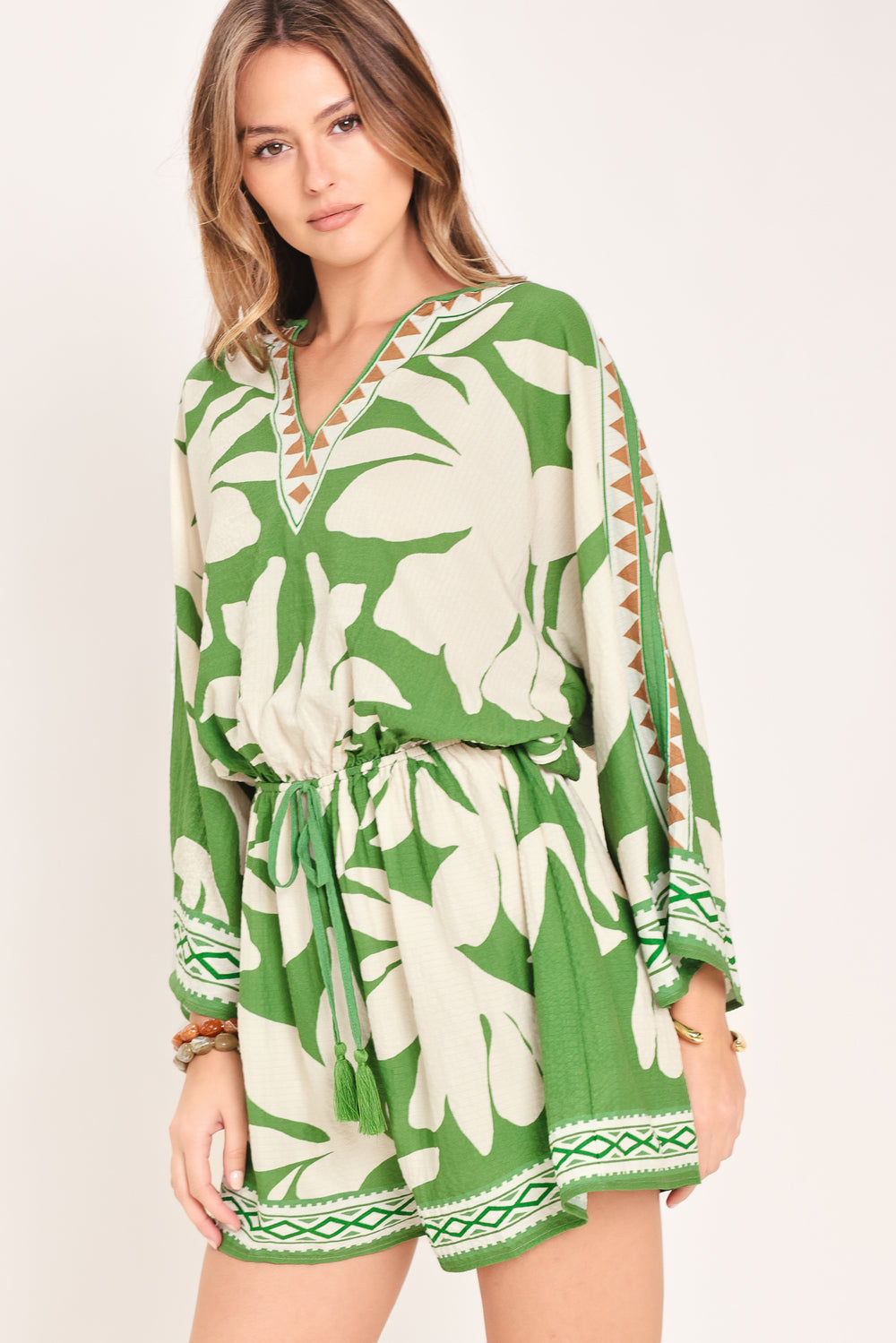 Woman wearing a green and white leaf pattern romper featuring V neckline, kimono sleeve, elastic waist with tie tassel and back neck button closure | Flying Tomato