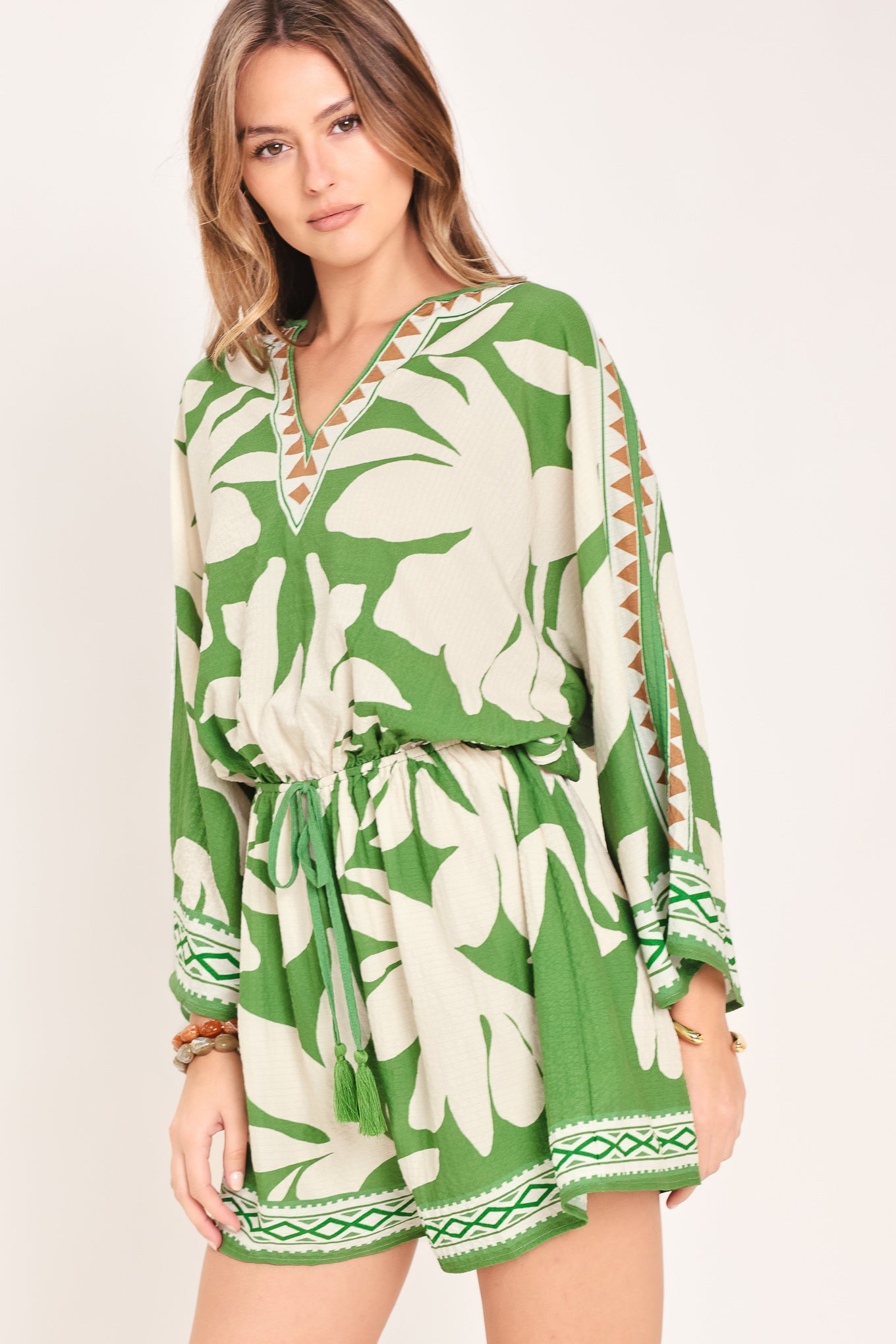 Woman wearing a green and white leaf pattern romper featuring V neckline, kimono sleeve, elastic waist with tie tassel and back neck button closure | Flying Tomato