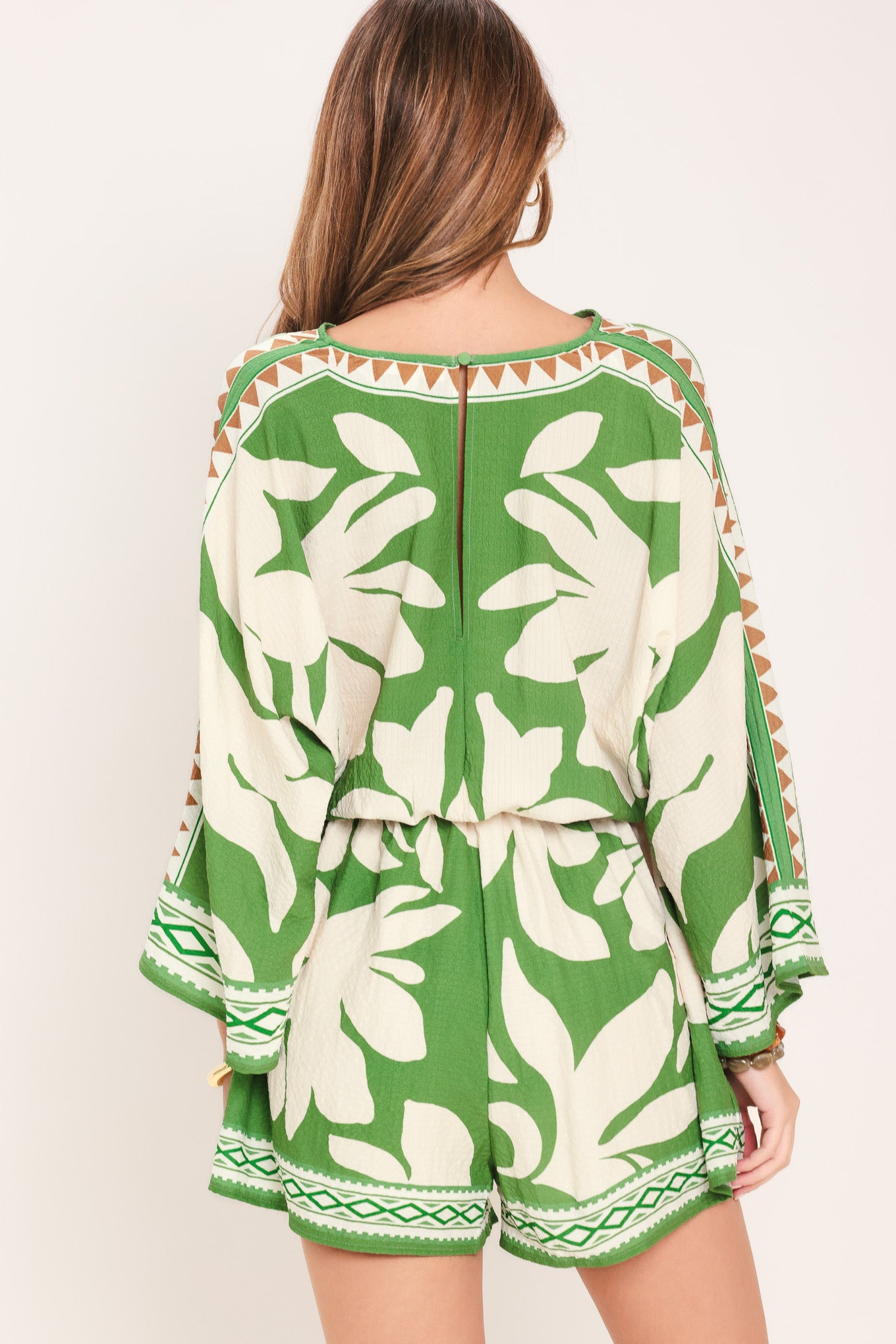 Woman wearing a green and white leaf pattern romper featuring V neckline, kimono sleeve, elastic waist with tie tassel and back neck button closure | Flying Tomato