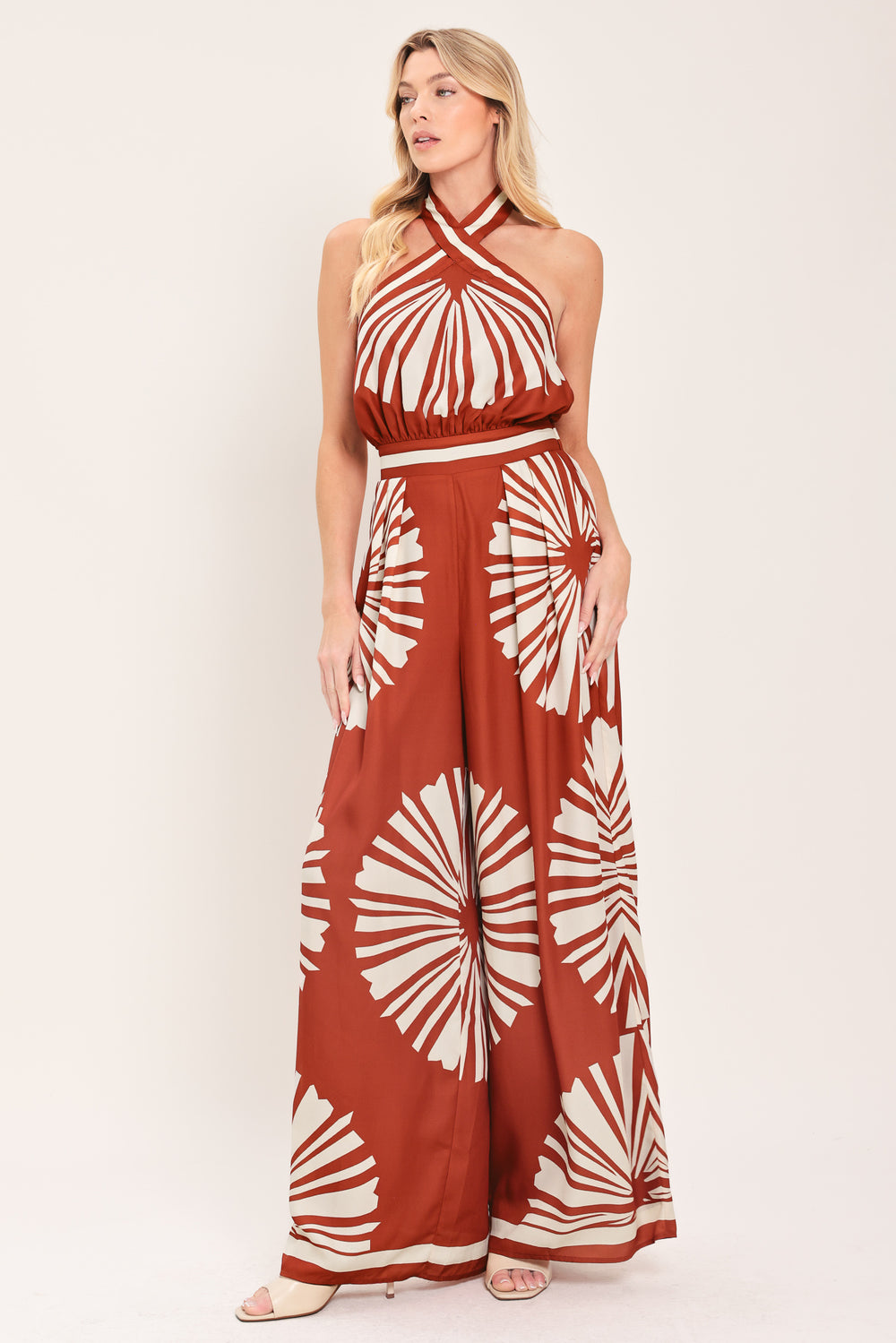 Woman wearing a terracotta wide leg jumpsuit with beige circular print featuring halter neckline with twist strap and back button closure, and bare back with tie closure | Flying Tomato