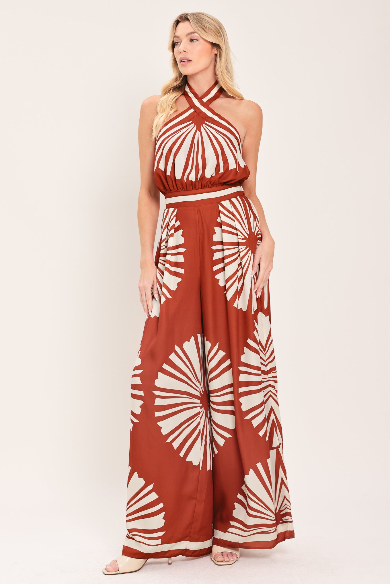 Woman wearing a terracotta wide leg jumpsuit with beige circular print featuring halter neckline with twist strap and back button closure, and bare back with tie closure | Flying Tomato