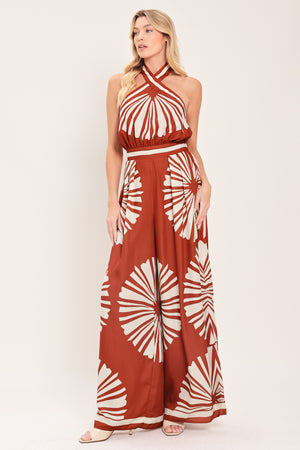 Woman wearing a terracotta wide leg jumpsuit with beige circular print featuring halter neckline with twist strap and back button closure, and bare back with tie closure | Flying Tomato