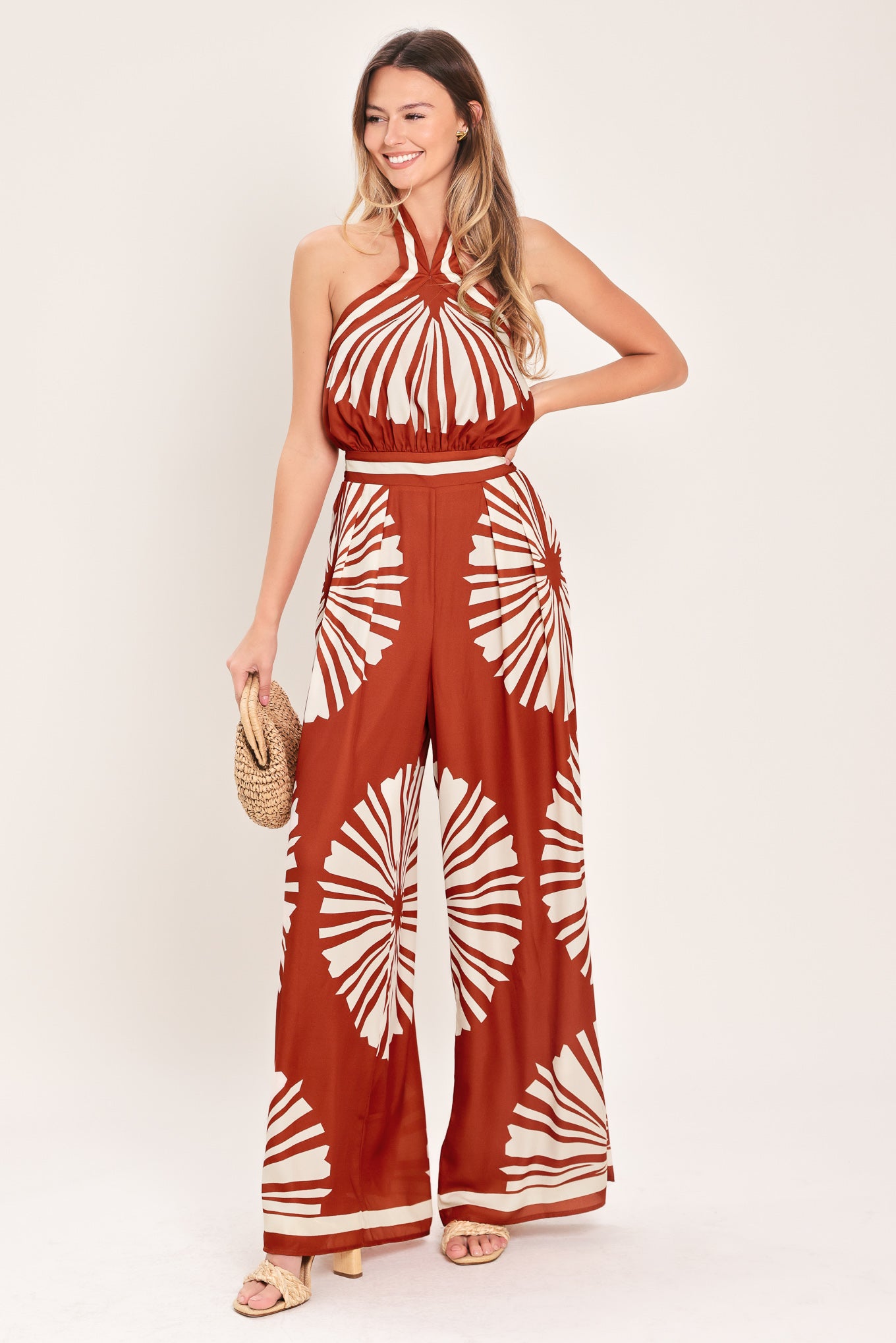 Woman wearing a terracotta wide leg jumpsuit with beige circular print featuring halter neckline with twist strap and back button closure, and bare back with tie closure | Flying Tomato