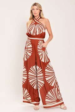 Woman wearing a terracotta wide leg jumpsuit with beige circular print featuring halter neckline with twist strap and back button closure, and bare back with tie closure | Flying Tomato