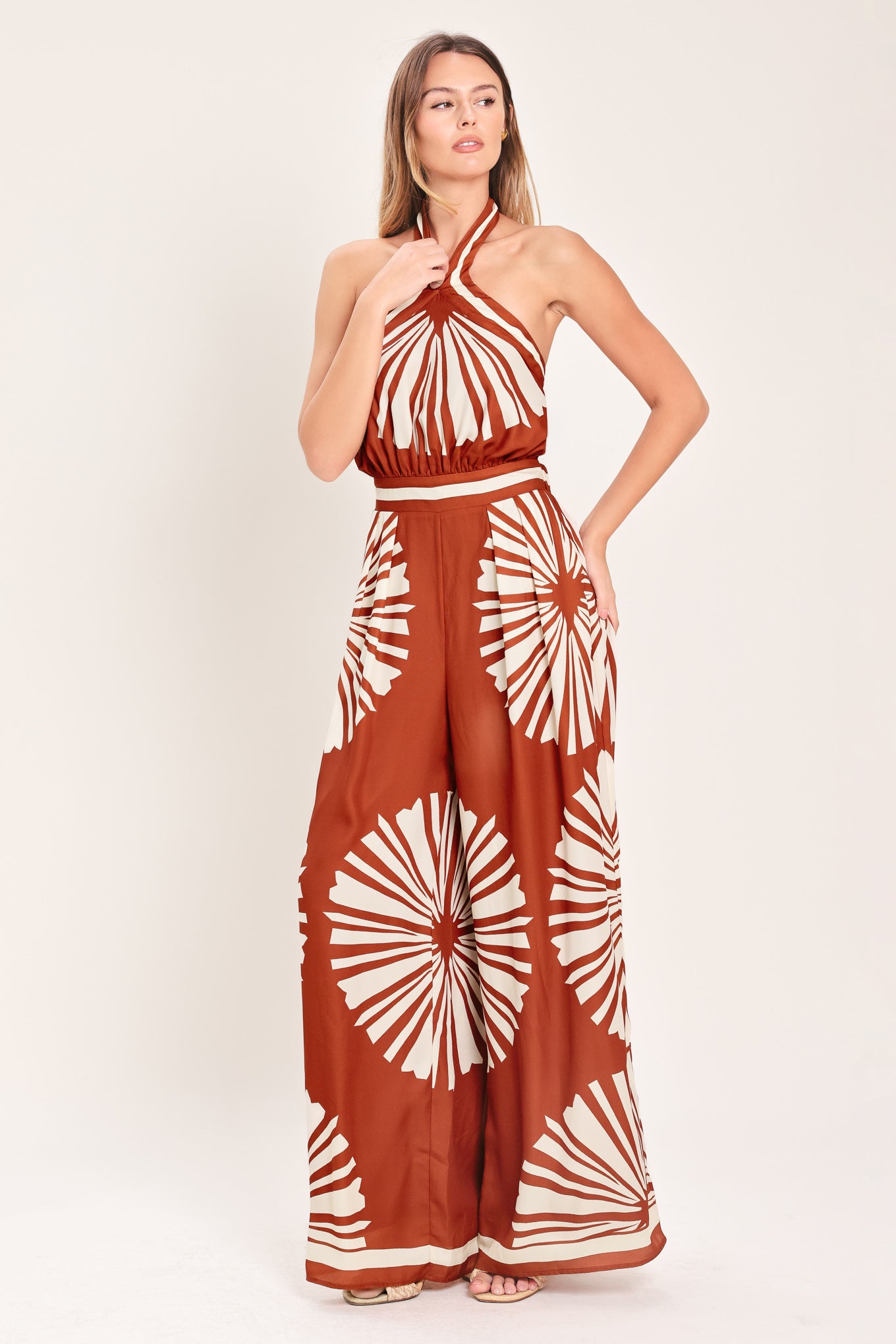 Woman wearing a terracotta wide leg jumpsuit with beige circular print featuring halter neckline with twist strap and back button closure, and bare back with tie closure | Flying Tomato