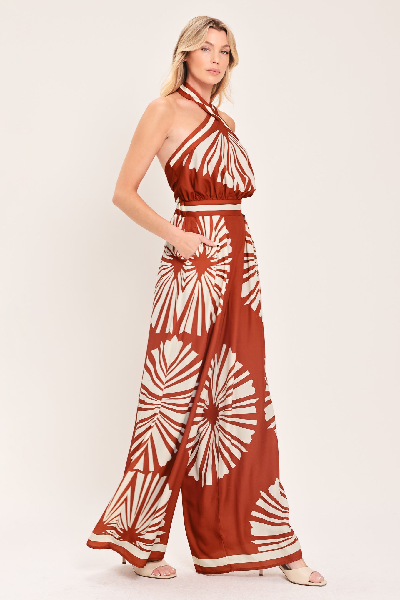 Woman wearing a terracotta wide leg jumpsuit with beige circular print featuring halter neckline with twist strap and back button closure, and bare back with tie closure | Flying Tomato
