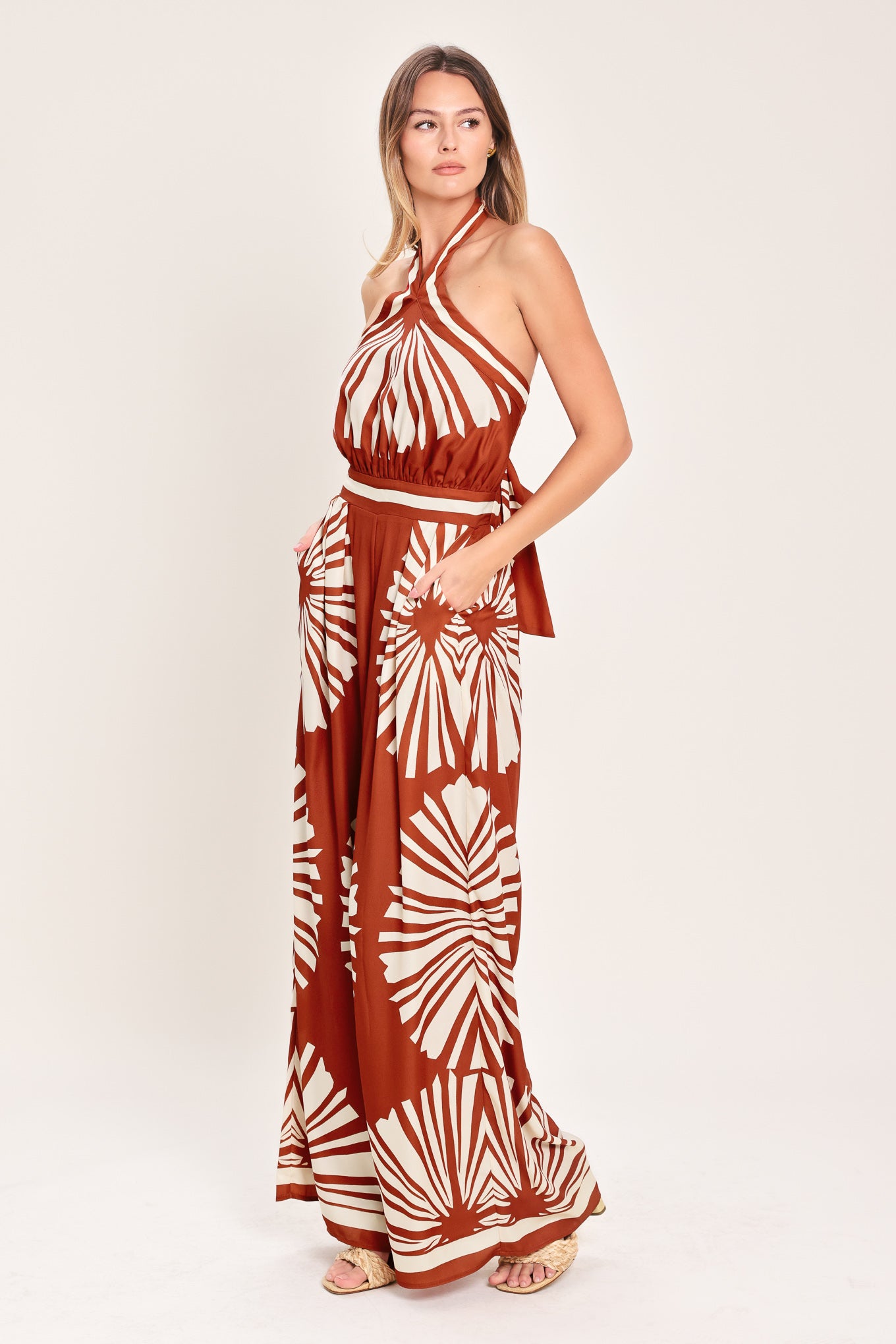 Woman wearing a terracotta wide leg jumpsuit with beige circular print featuring halter neckline with twist strap and back button closure, and bare back with tie closure | Flying Tomato
