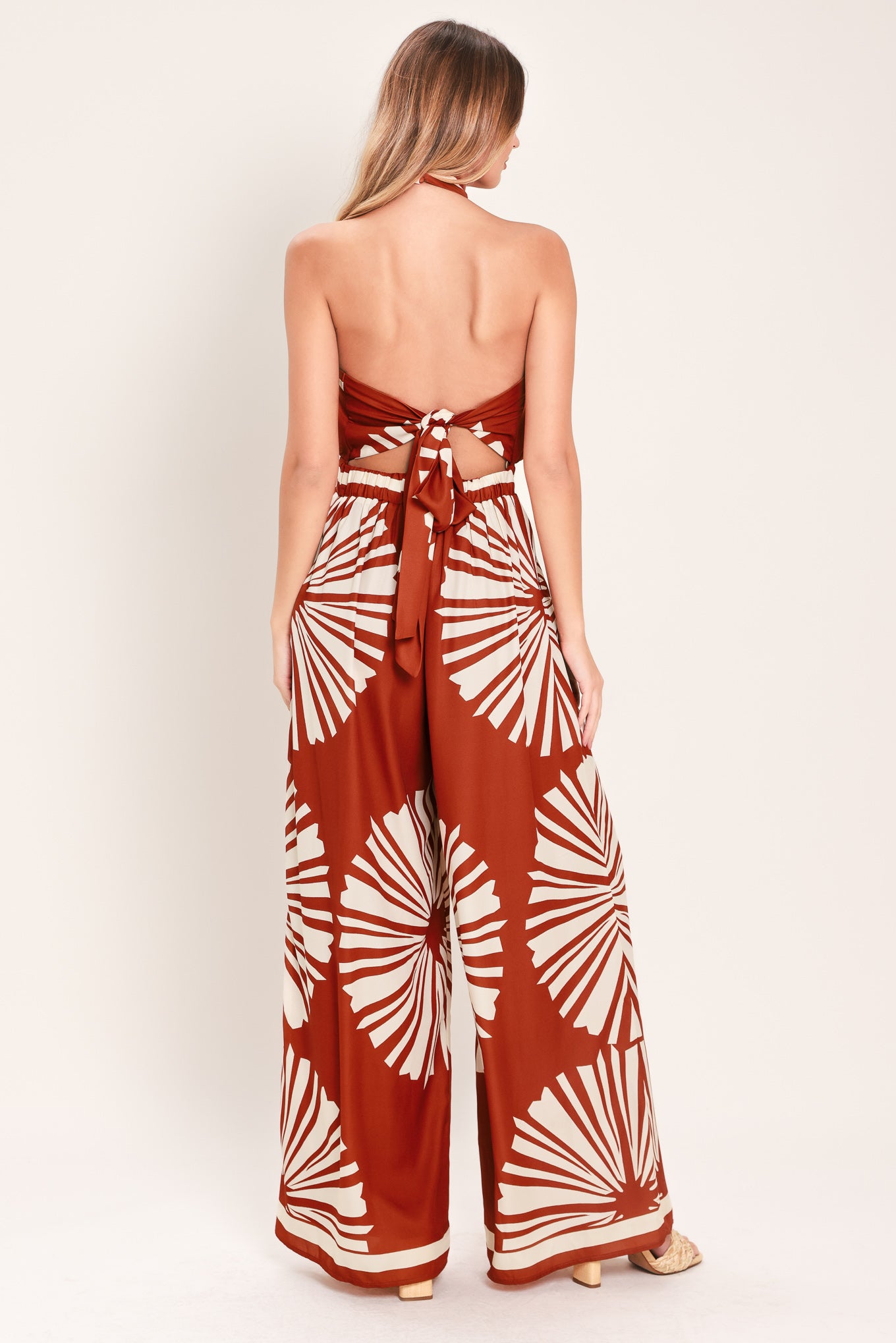 Woman wearing a terracotta wide leg jumpsuit with beige circular print featuring halter neckline with twist strap and back button closure, and bare back with tie closure | Flying Tomato