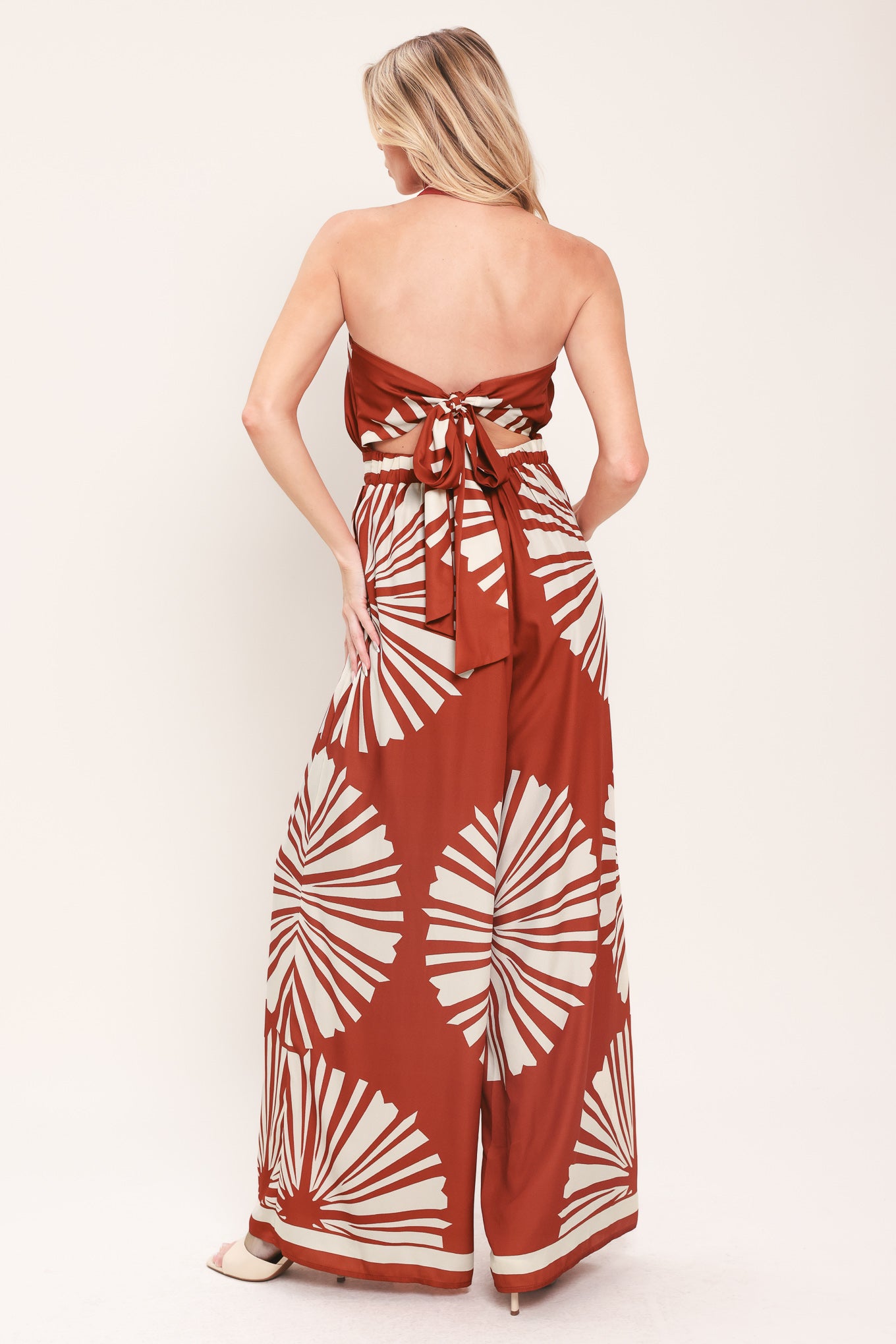 Woman wearing a terracotta wide leg jumpsuit with beige circular print featuring halter neckline with twist strap and back button closure, and bare back with tie closure | Flying Tomato