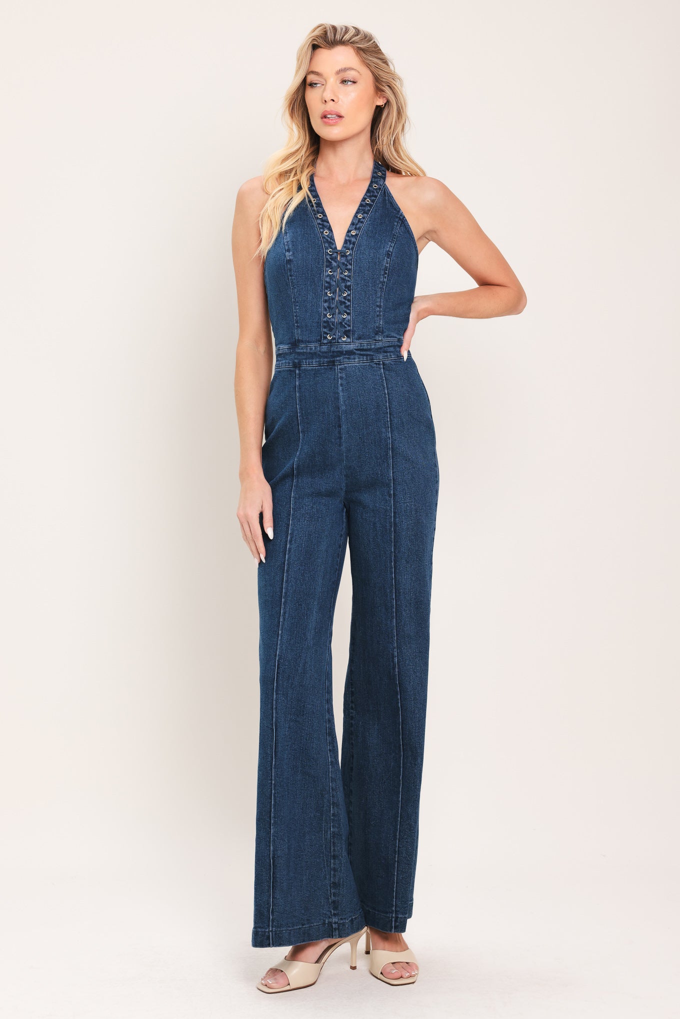 Woman wearing a denim jumpsuit featuring halter neckline with eyelet placket, boot cut leg and bare back with zipper closure | Flying Tomato