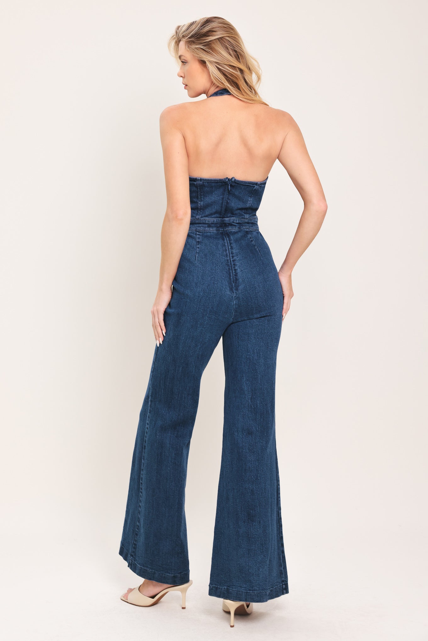 Woman wearing a denim jumpsuit featuring halter neckline with eyelet placket, boot cut leg and bare back with zipper closure | Flying Tomato