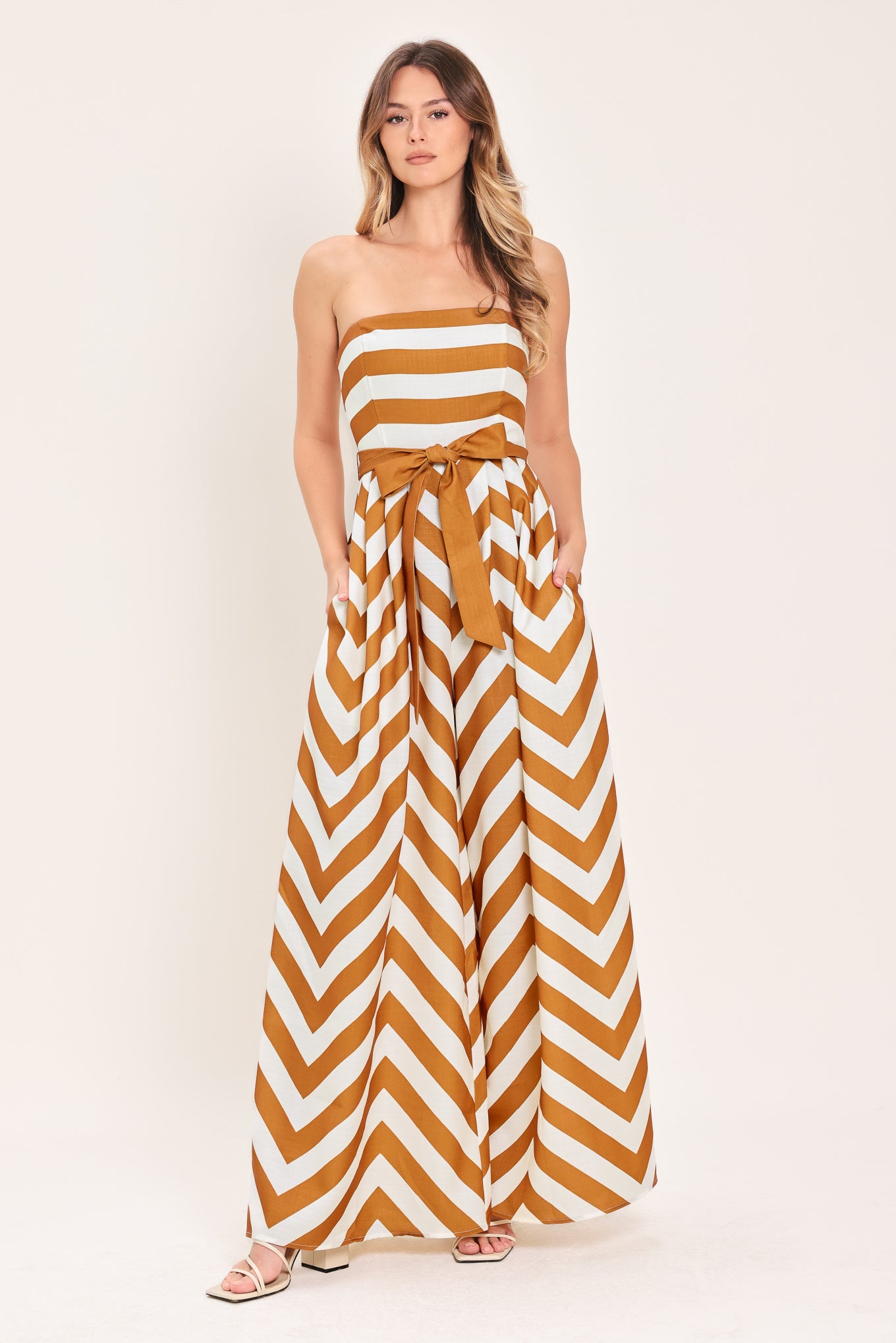 Woman wearing a camel and white strapless, long chevron-patterned jumpsuit featuring solid tie, wide leg and smocked back bodice | Flying Tomato