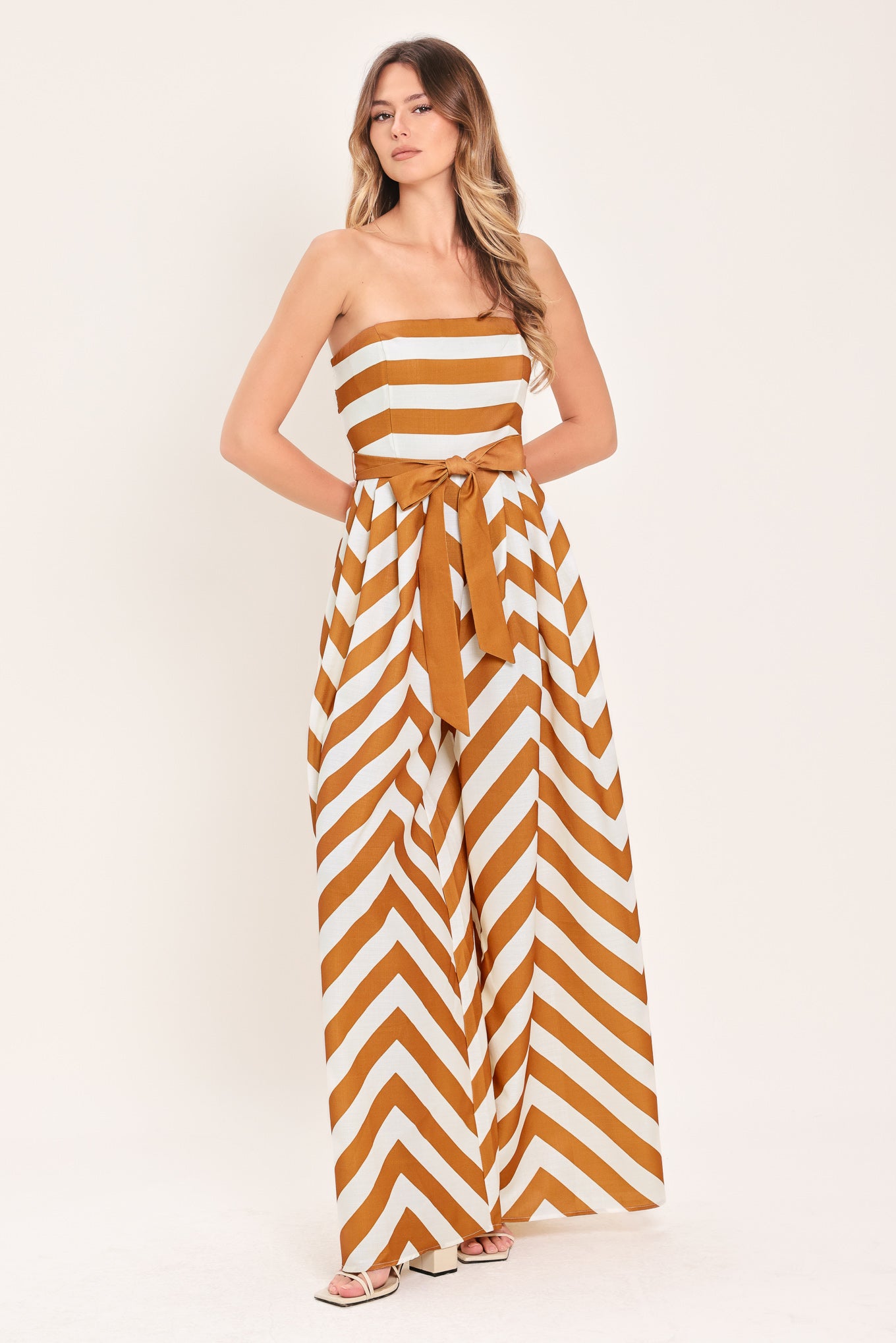 Woman wearing a camel and white strapless, long chevron-patterned jumpsuit featuring solid tie, wide leg and smocked back bodice | Flying Tomato