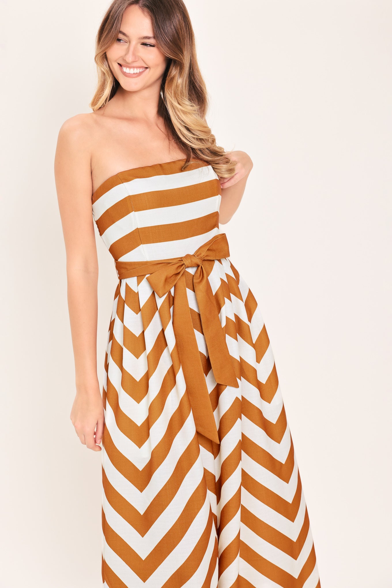 Woman wearing a camel and white strapless, long chevron-patterned jumpsuit featuring solid tie, wide leg and smocked back bodice | Flying Tomato