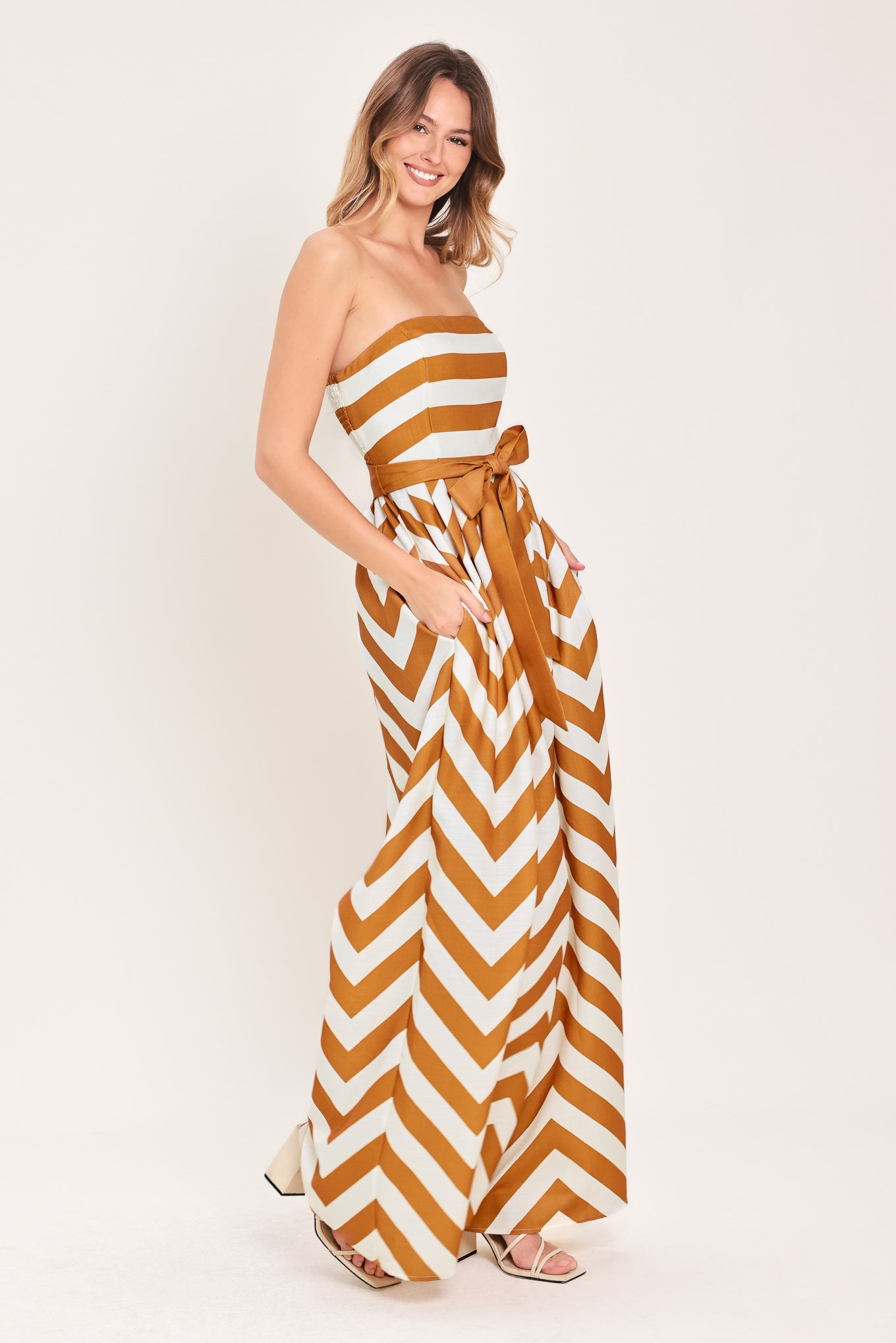 Woman wearing a camel and white strapless, long chevron-patterned jumpsuit featuring solid tie, wide leg and smocked back bodice | Flying Tomato